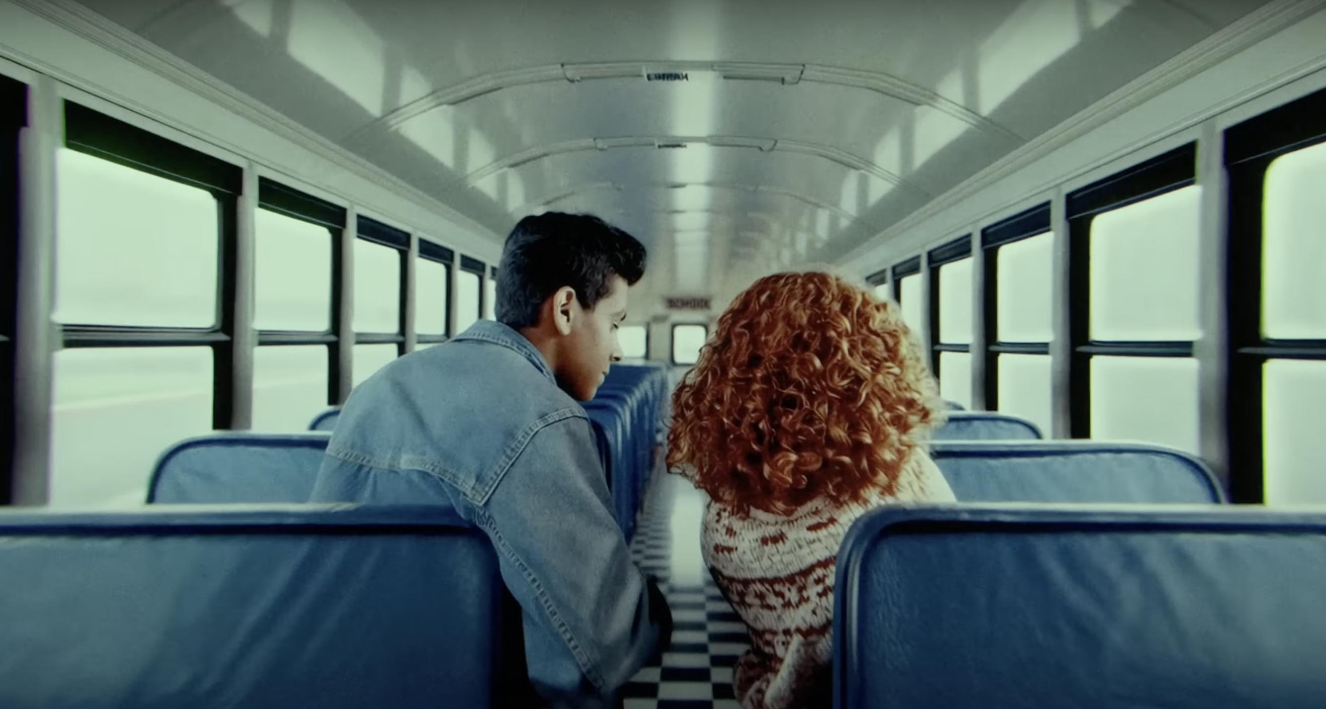 The inside of a school bus. Two students, a brown-skinned boy with a cropped hair cut and a girl with big red curly hair, are seen from the back. 