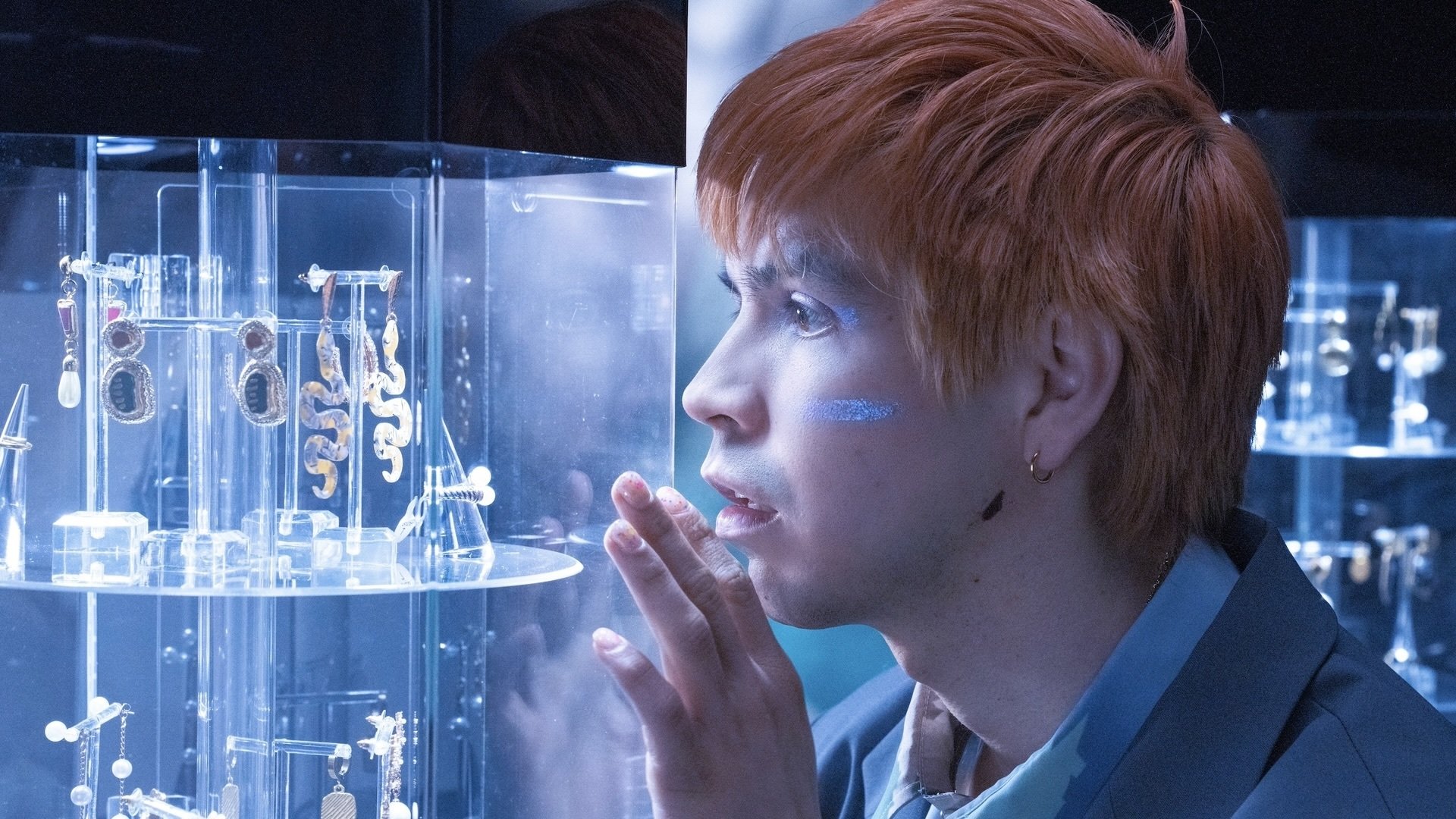 A man with orange hair admires a golden oyster earring in a jewelry display case.