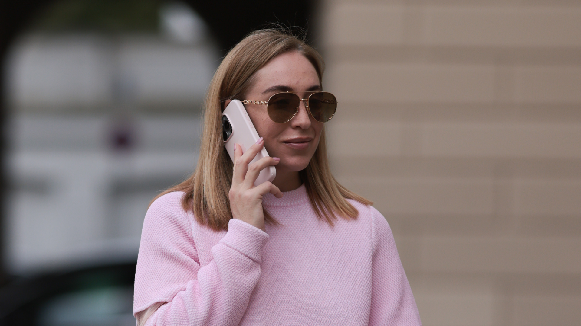A woman wearing a pink sweater and sunglasses holds a phone to her ear.