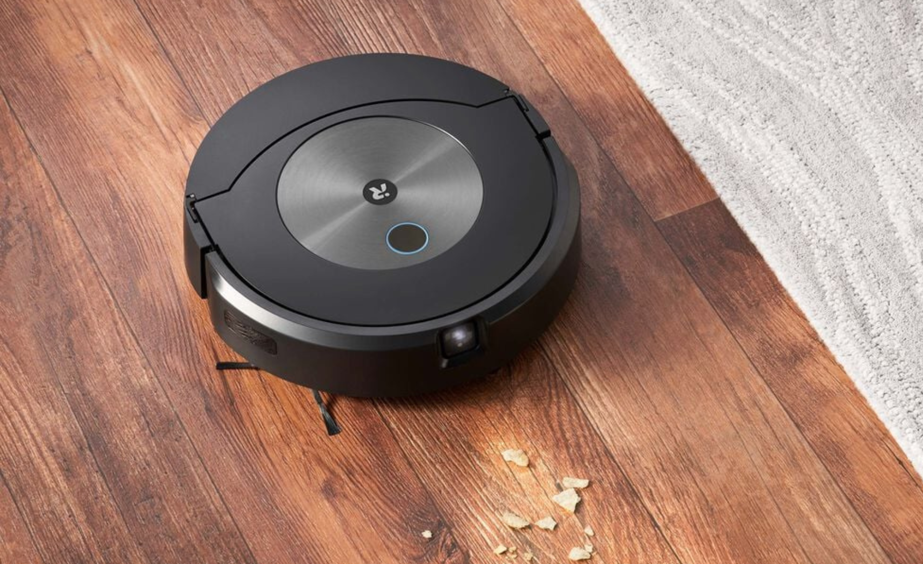 Roomba robot vacuum cleaning crumbs on hardwood floor with rug in peripheral