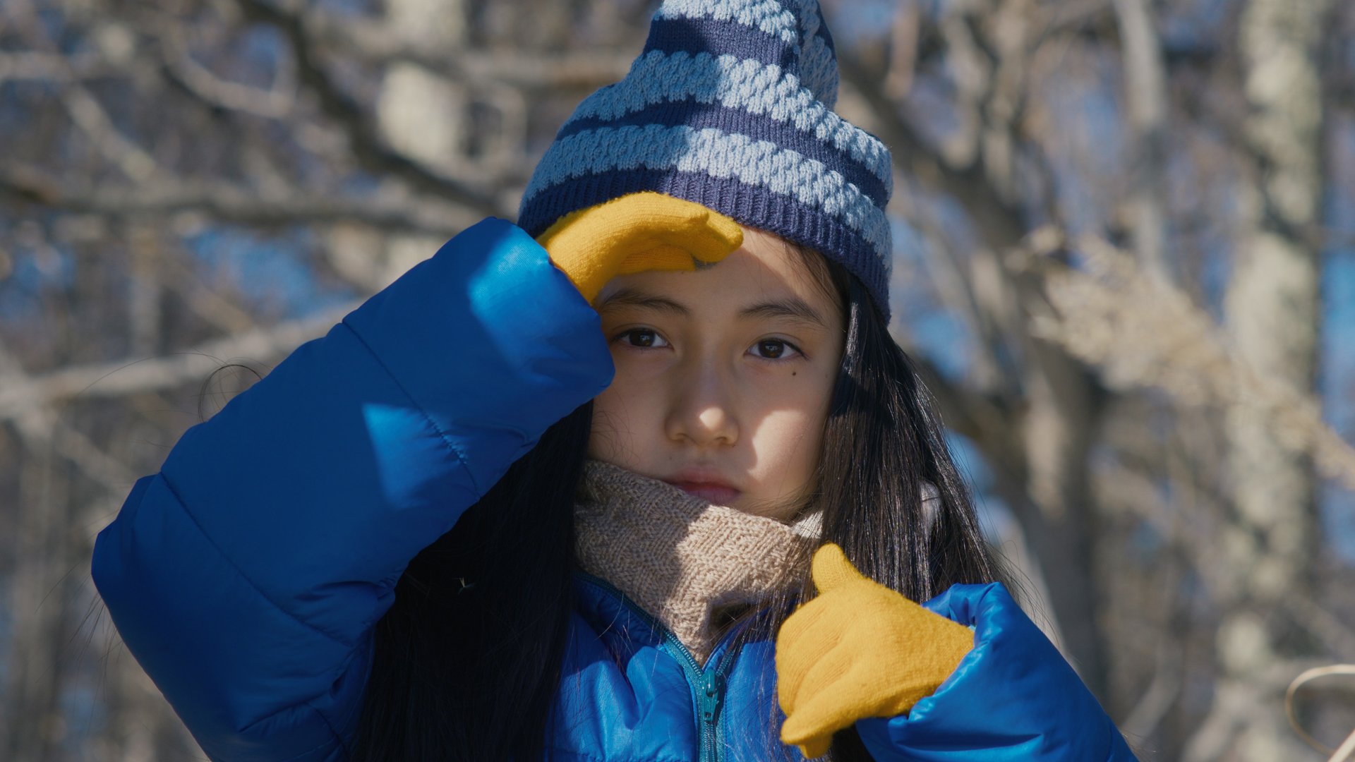 A young girl in bright winter clothing faces the camera, shielding her eyes from the sun.