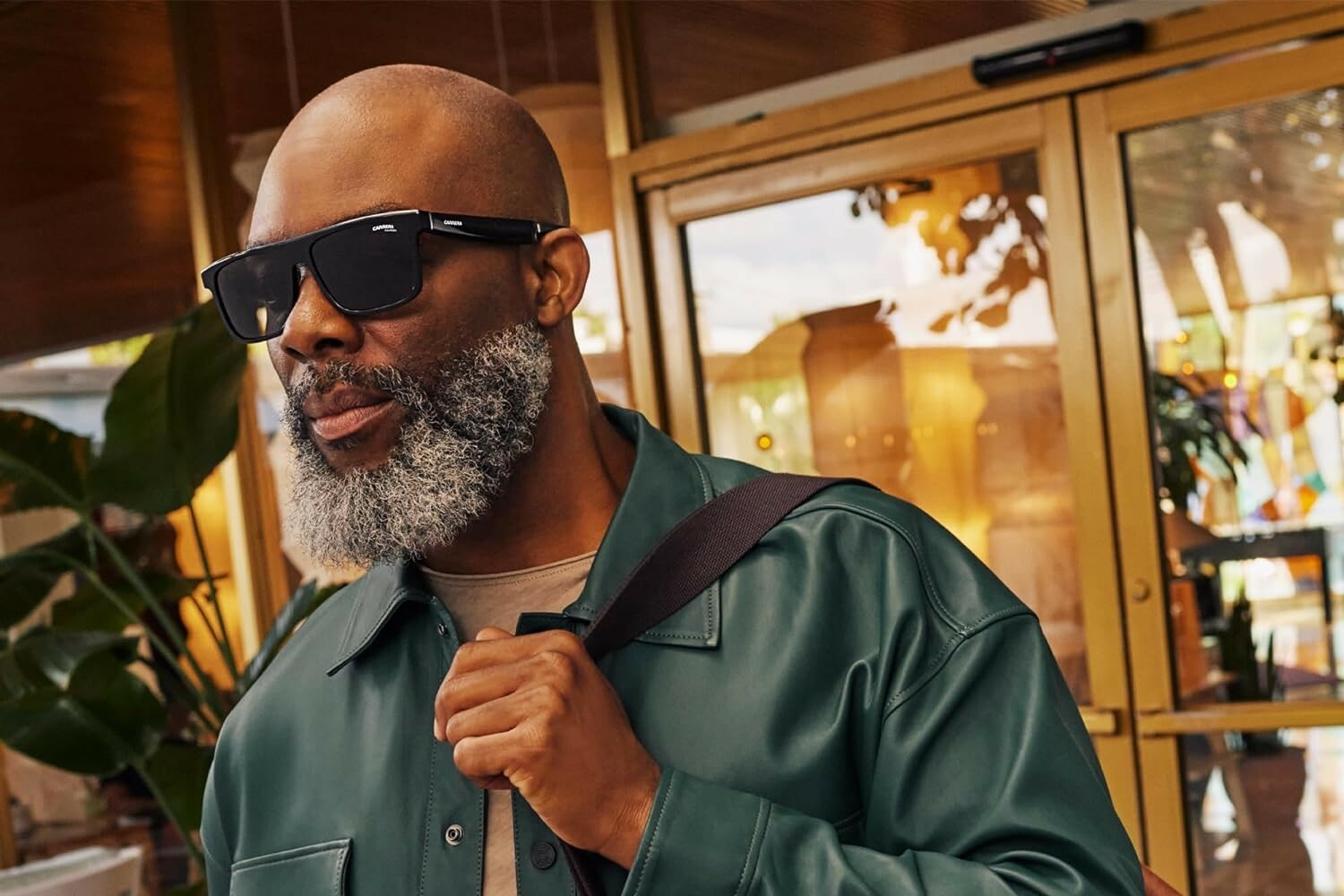 a person wearing the Carrera Smart Glasses with Alexa sunglasses holds a brown leather bag over their left shoulder while looking forward. the person has a salt and pepper beard and a bald head 