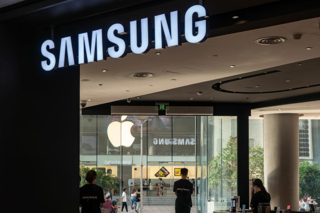 The Samsung logo over a retail store with the Apple logo visible through the glass doors beneath it