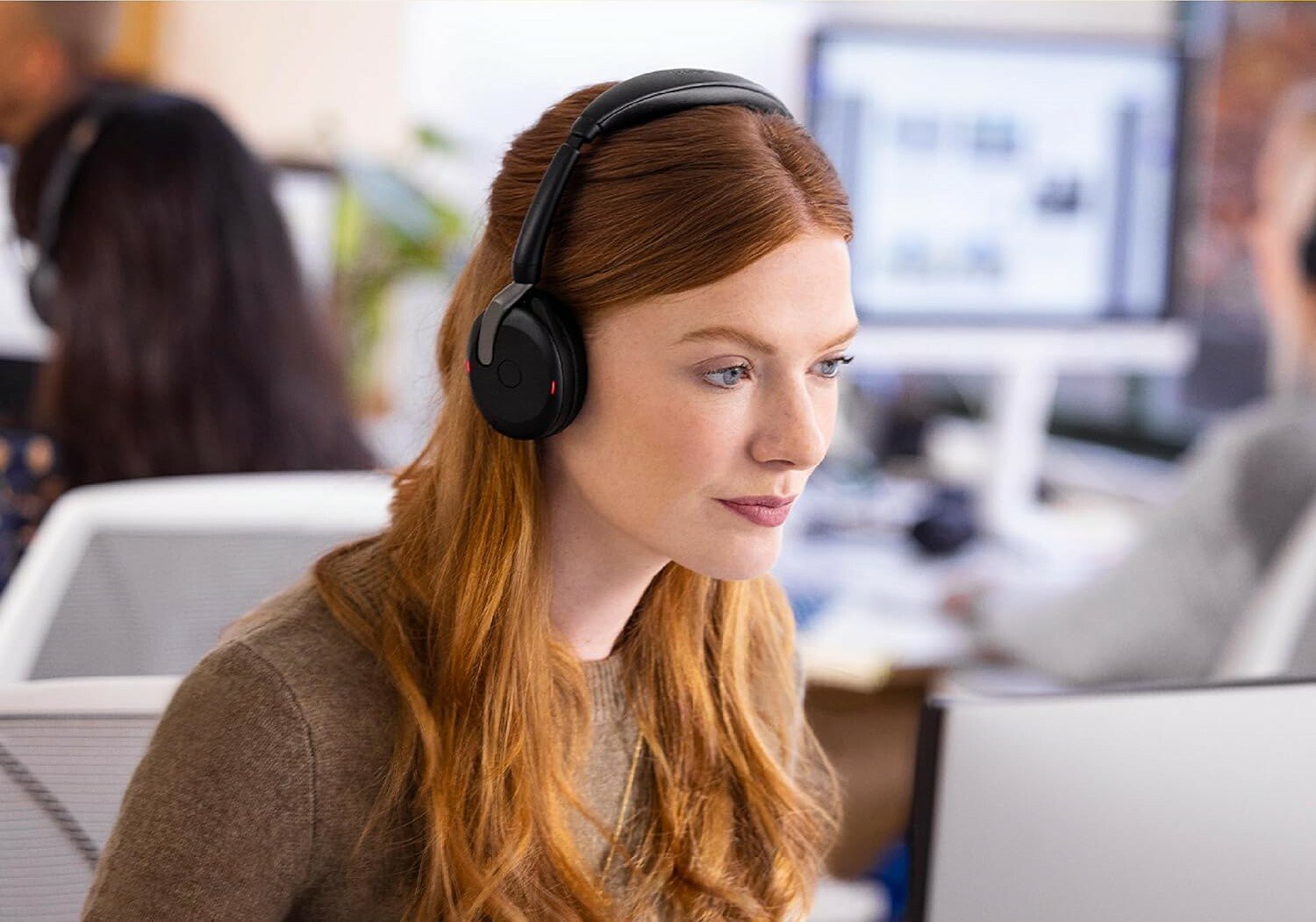 a person with red hair wears the  Jabra Evolve2 65 headset while looking at a computer screen. The setting it an office environment with other people working in the background at computer stations.