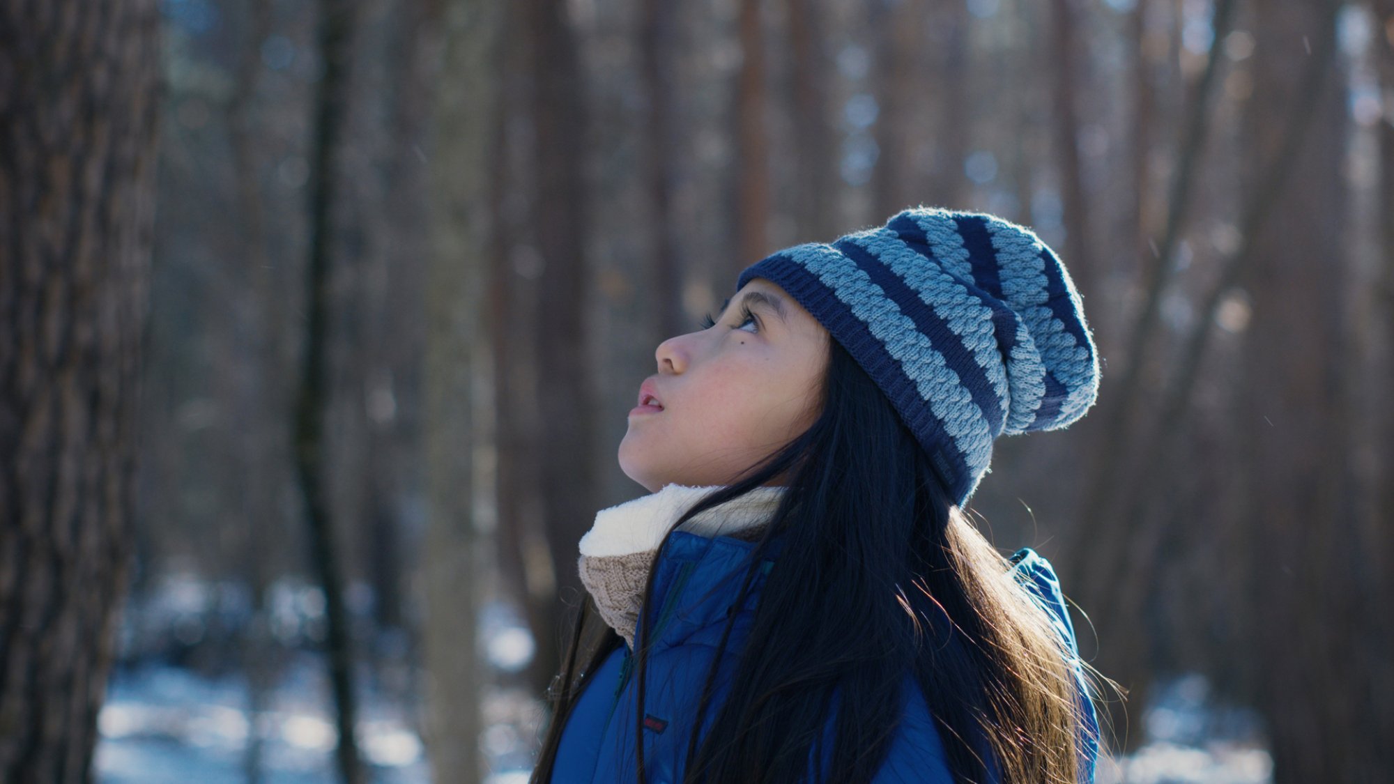 A young girl in winter clothing stands in a wood looking up at the sky.