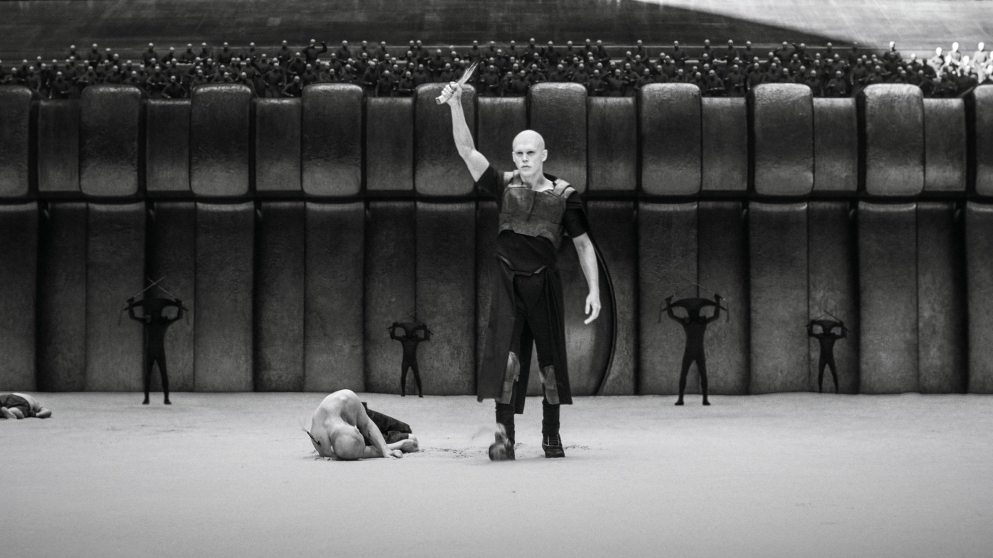 Feyd Rautha Harkonnen raises his sword while standing over a dead body in a crowded arena.