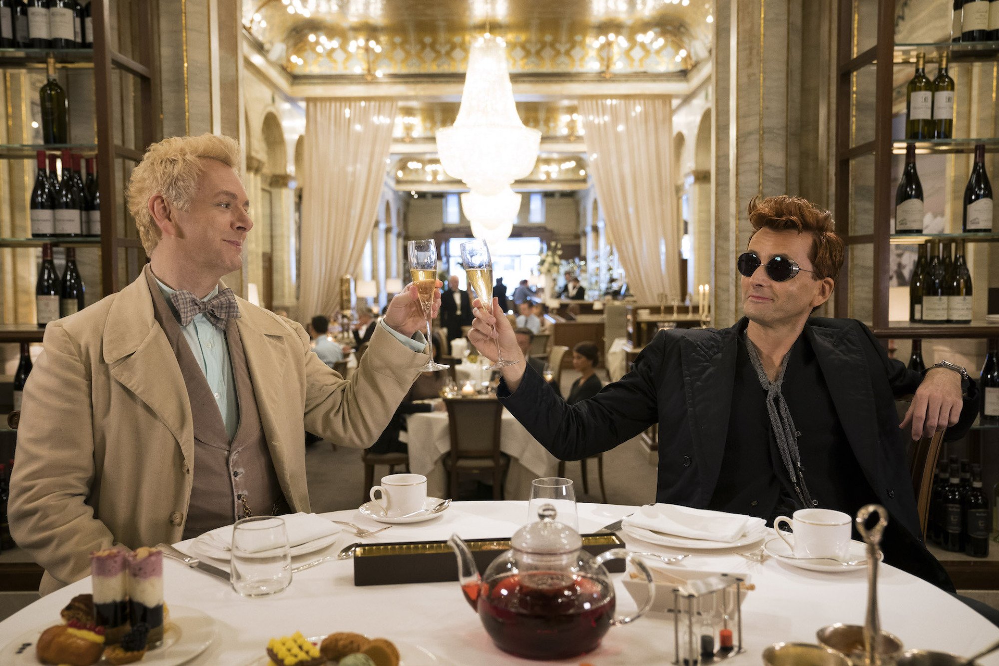 Two men, one in white/tan and one in black and sunglasses, clink glasses at a nice restaurant.