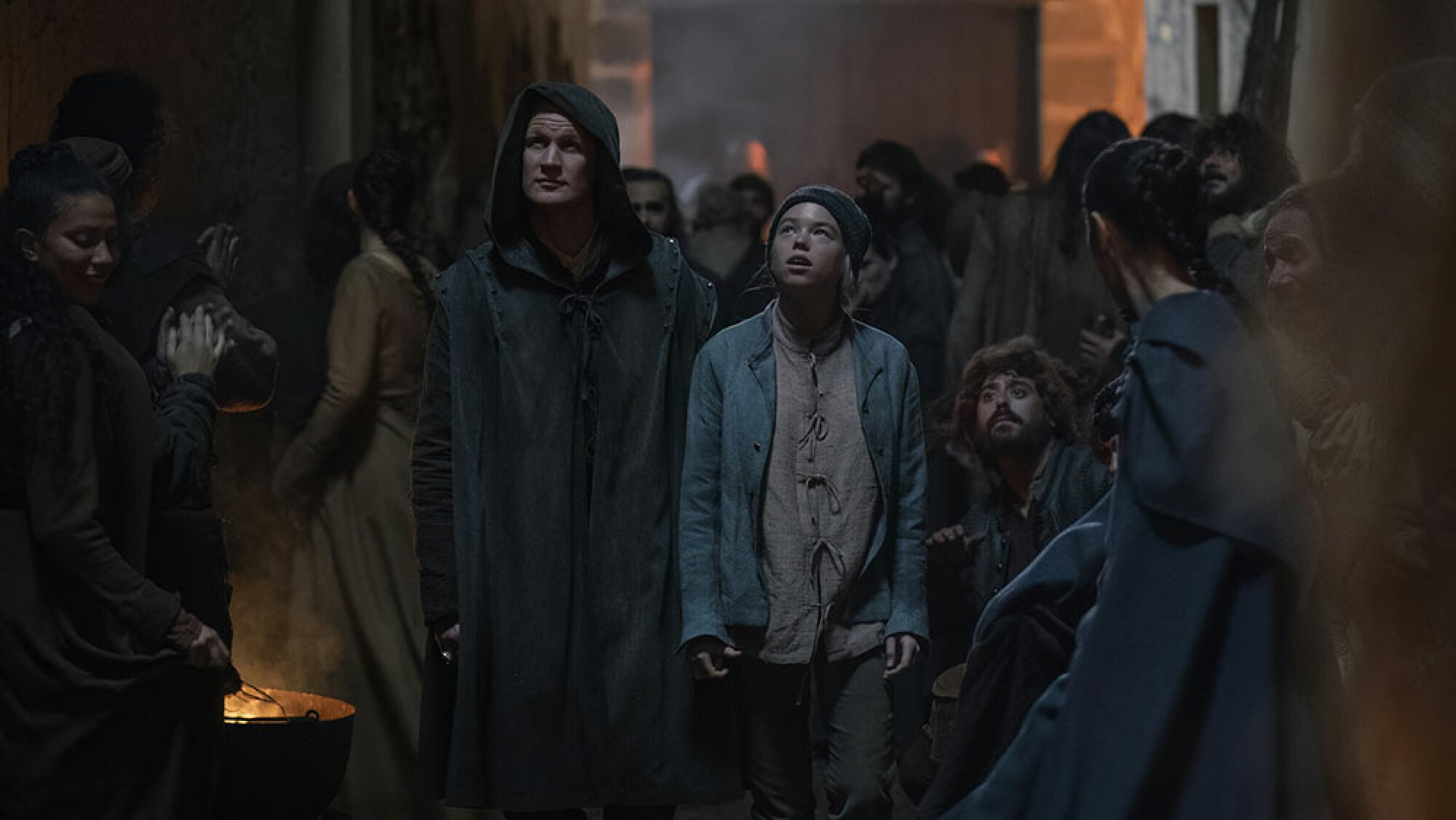 A man and a young woman walk through a busy street wearing cloaks.