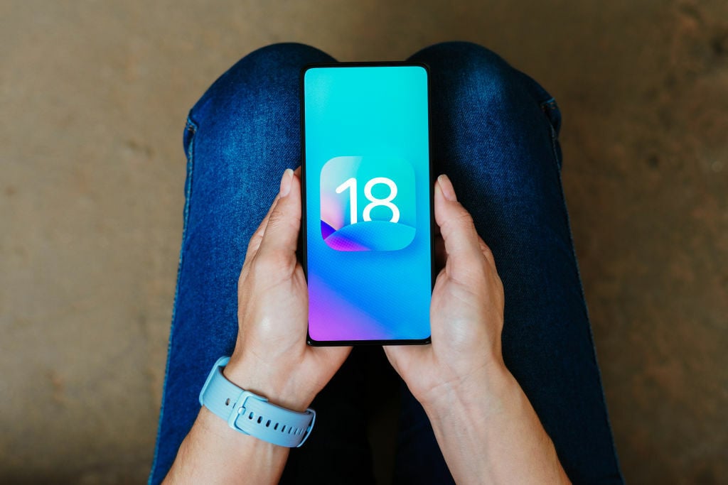 ios 18 logo on a smartphone being held in someone's hands on their lap