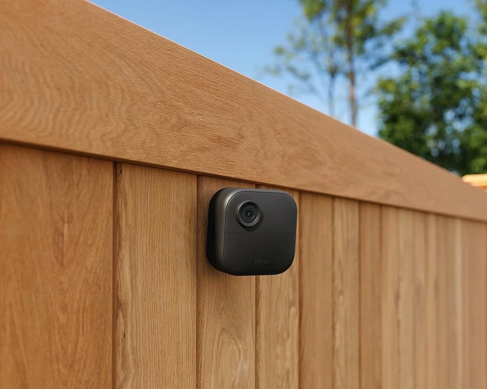 a blink outdoor 4 camera is mounted onto a wooden fence. Behind the fence is blue sky and green trees on a sunny day