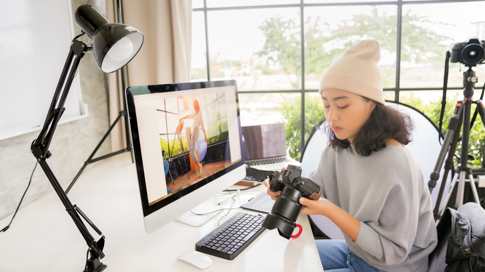 Side view of Asian female freelancer photographer cheking photos on a digital camera while sitting at the table in workstation - stock photo