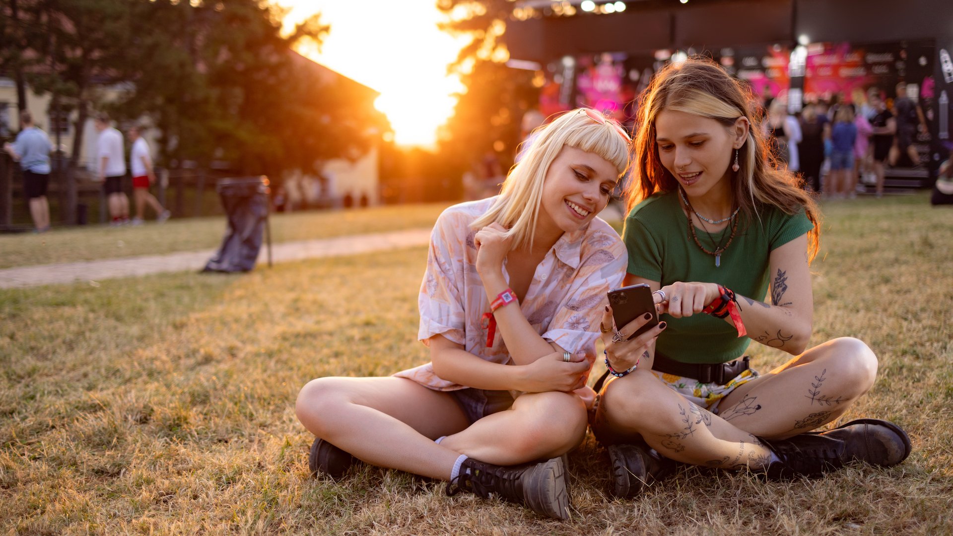 Friends reading a text message on cell phone while relaxing on grass during a music festival at sunset.