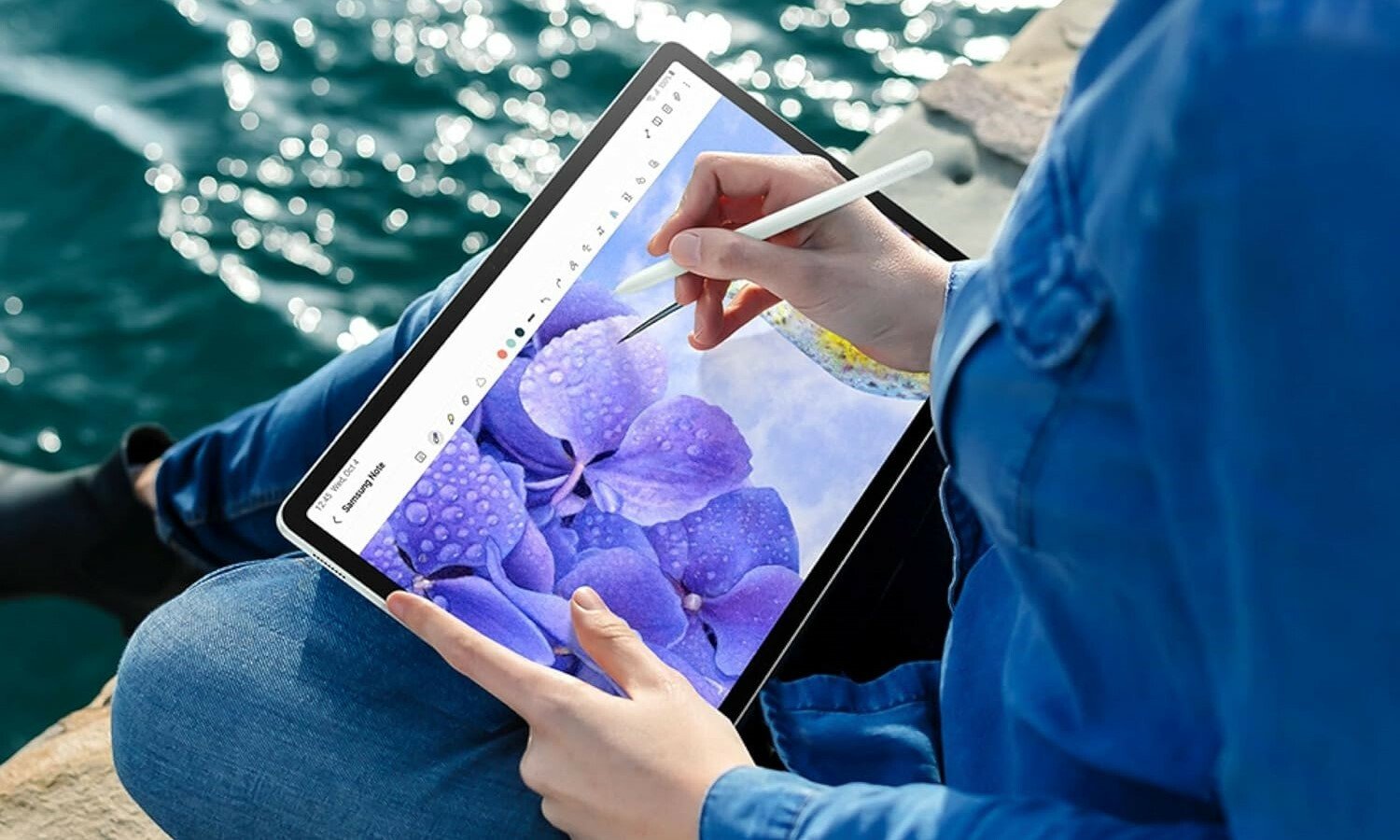 a person sits cross legged on some large rocks next to a body of water. They're using the included pen to draw on the  Samsung Galaxy Tab S9 FE+ tablet 