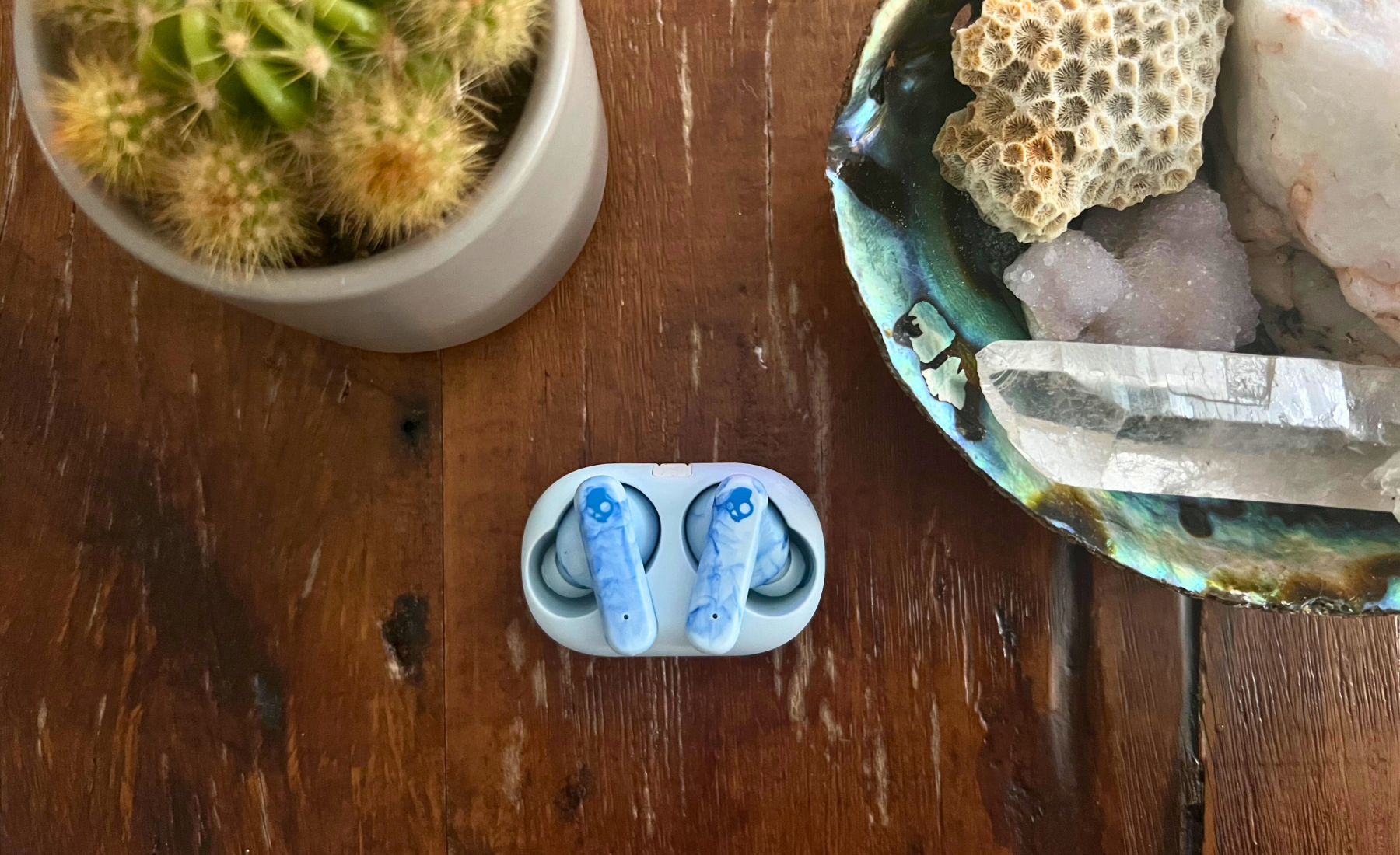 Skullcandy EcoBuds headphones in case laying on wooden coffee table with potted cactus and seashell decor in peripherals