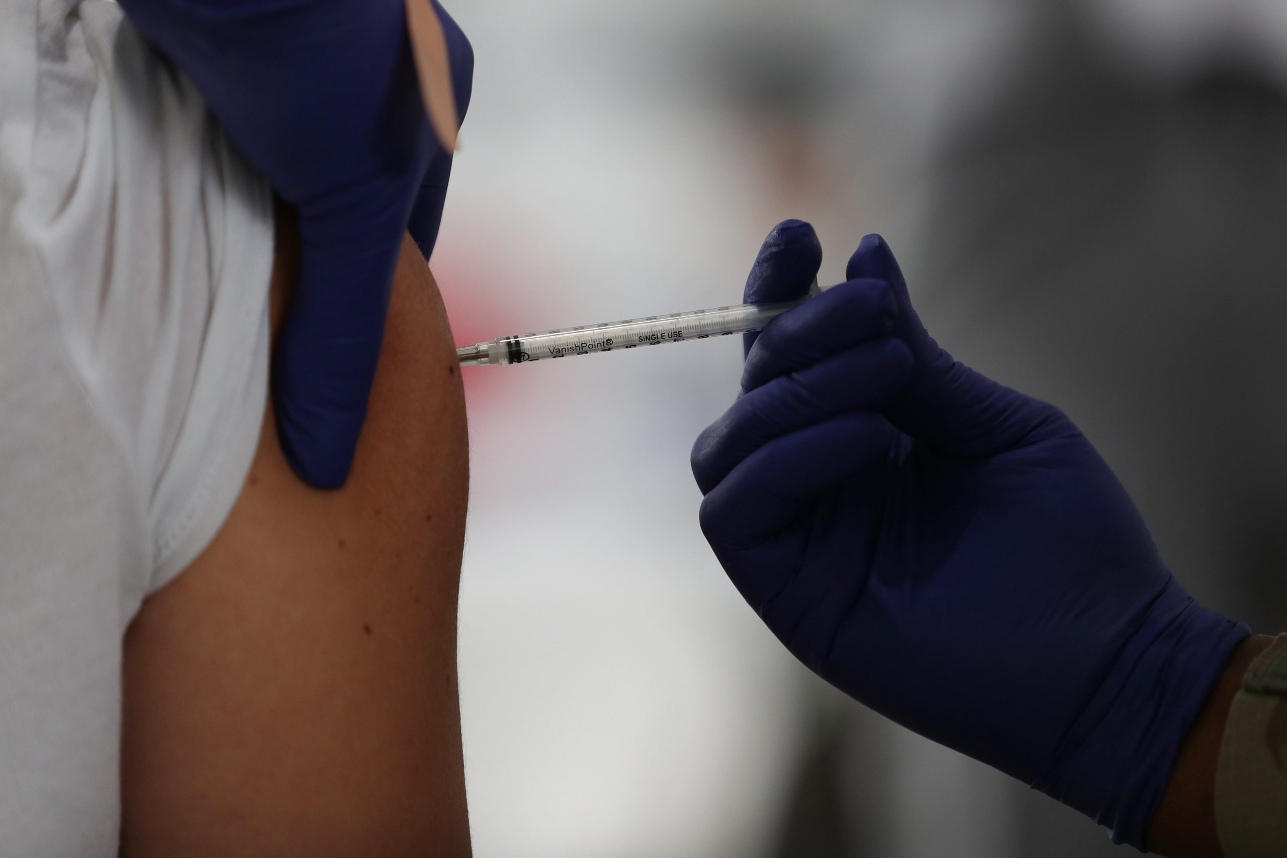 A gloved hand injects a needle into a person's upper arm.