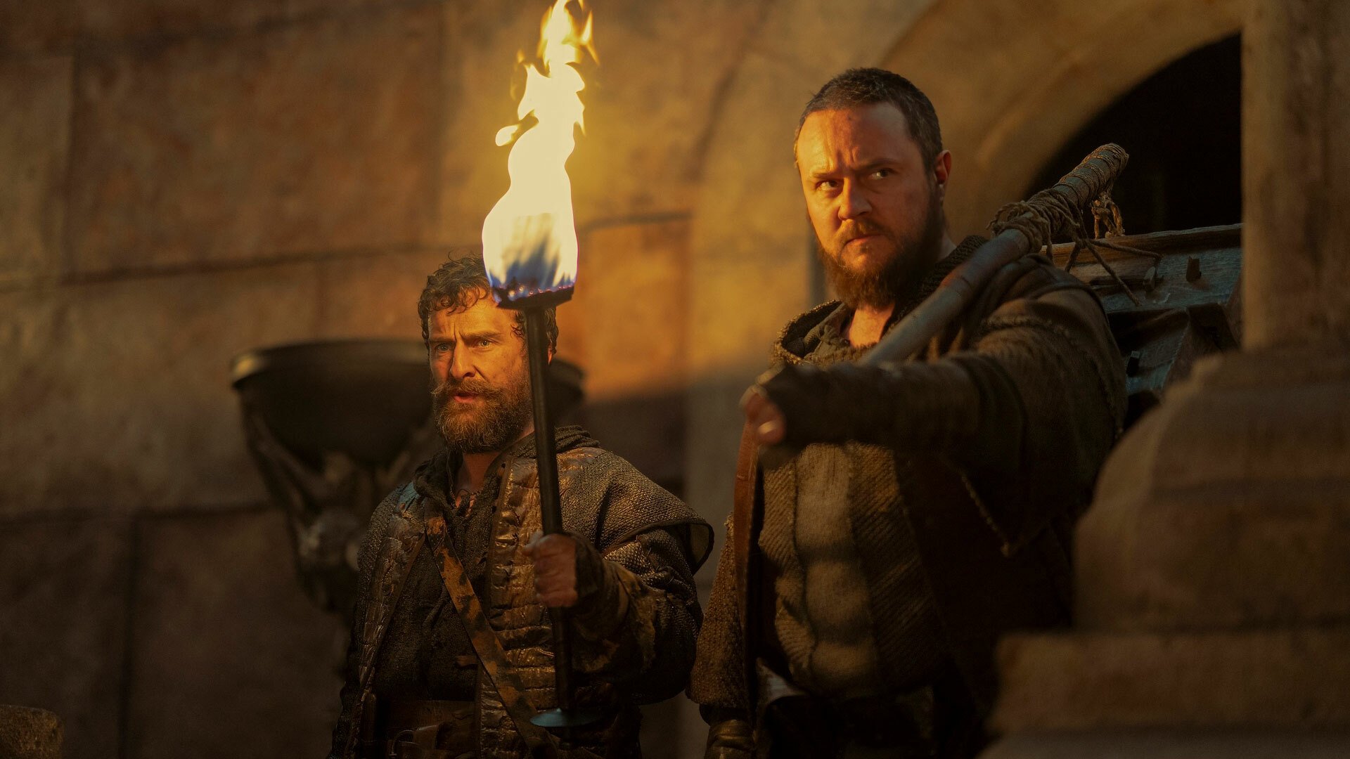 A man with a beard and a larger man stand in a castle holding a fiery torch.