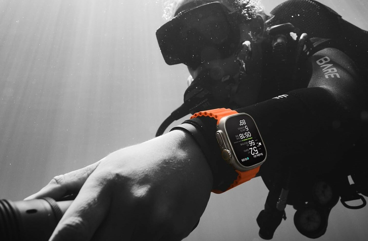 a person diving underwater wears the apple watch ultra 2 with an orange band. The photo is black and white aside from the orange band on the watch.