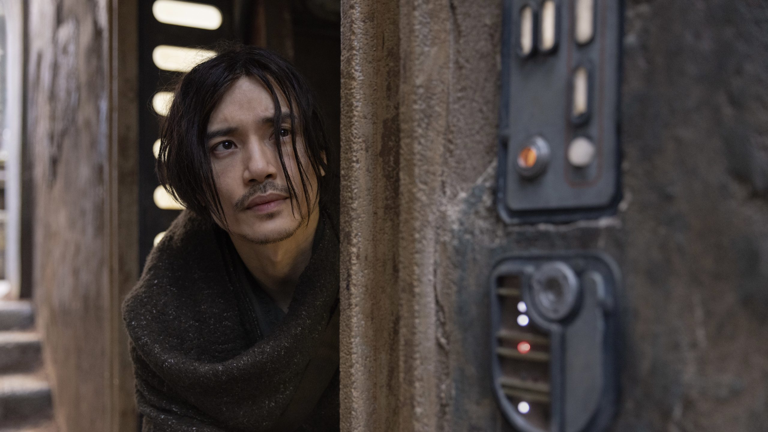 A man in a brown cloak peeks out of a doorway.