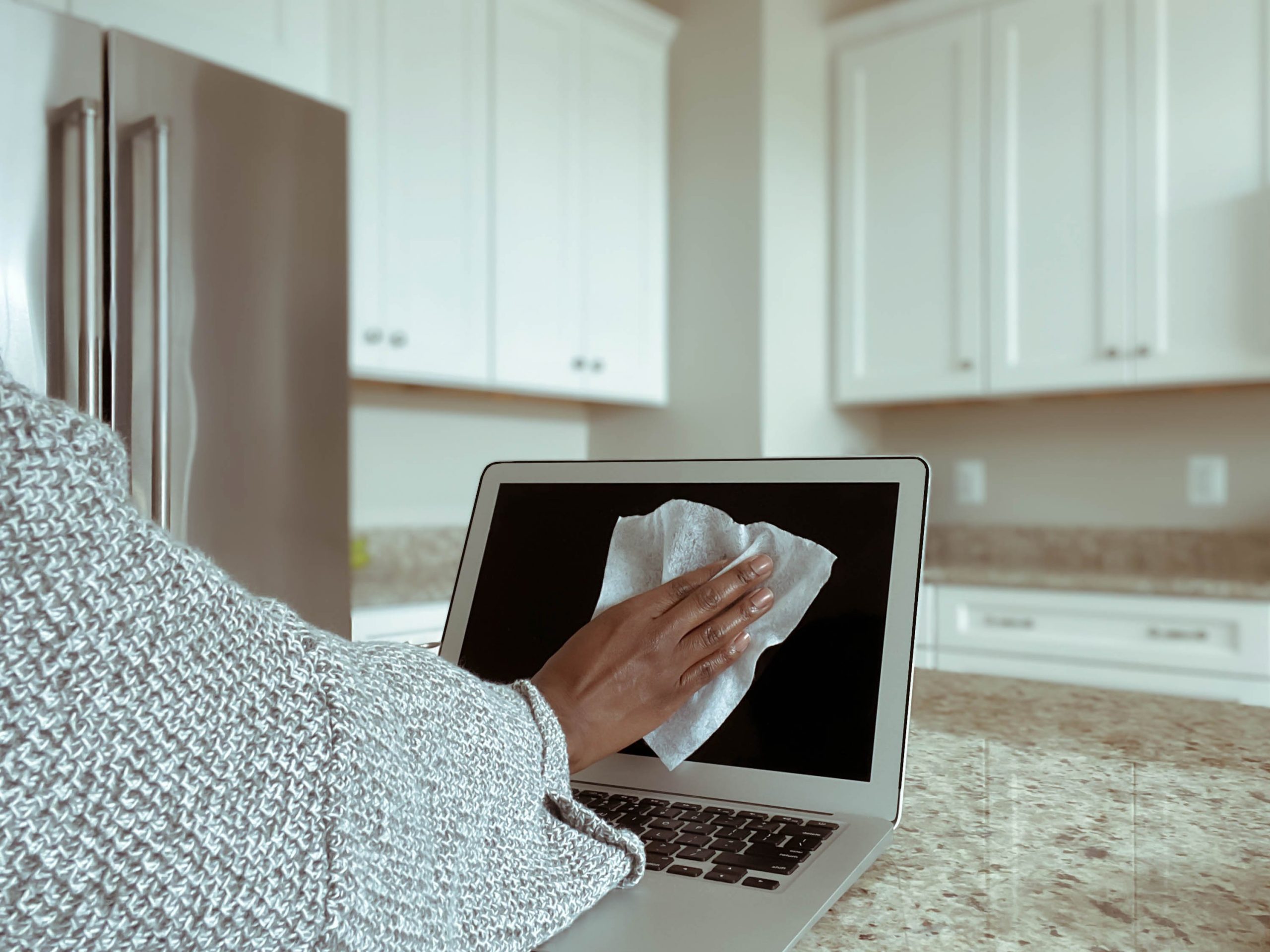 Close-up of unrecognizable woman in kitchen cleaning laptop screen with disinfectant wipe