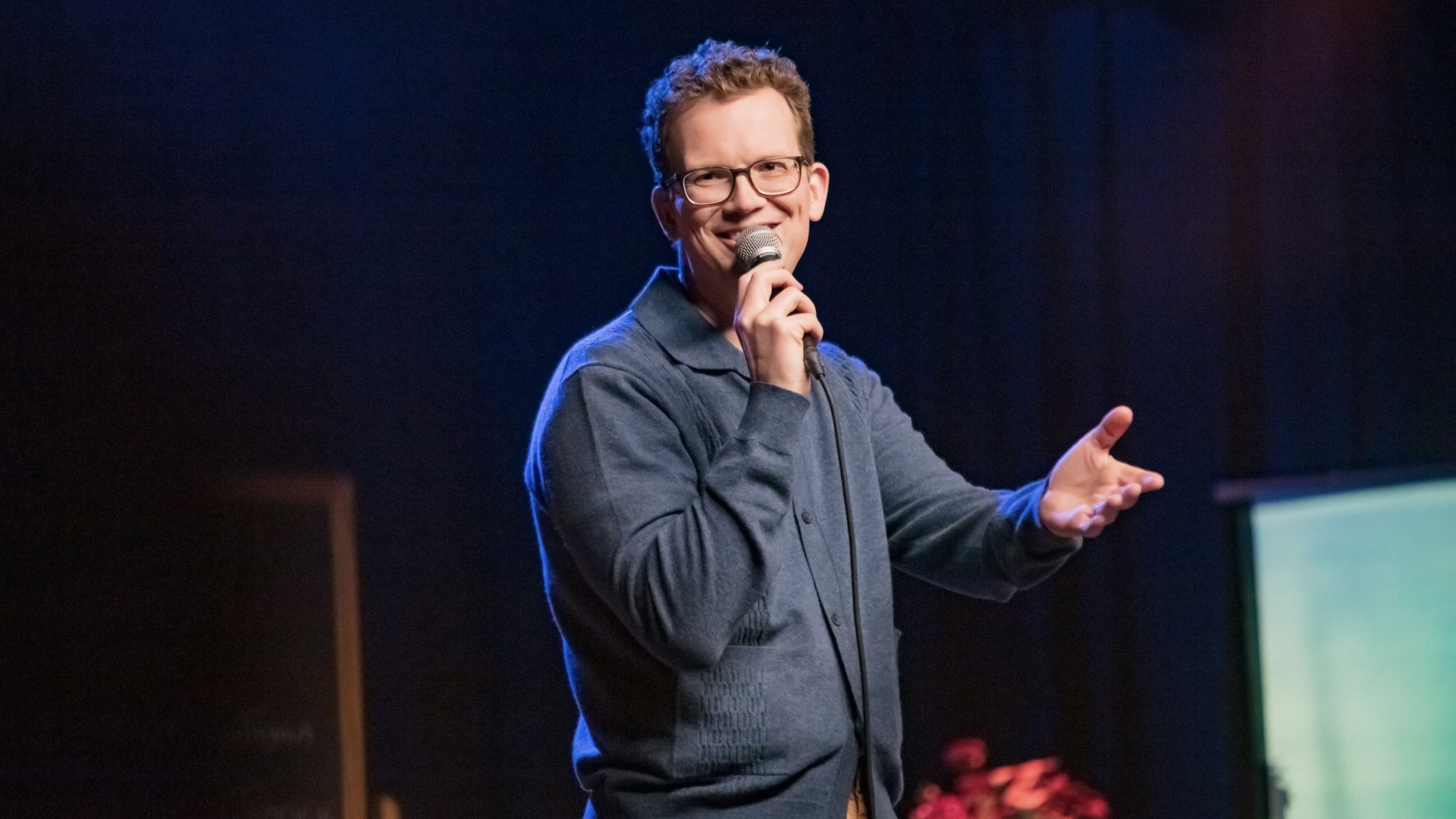 Hank Green performs stand-up onstage in a blue button-up sweater.