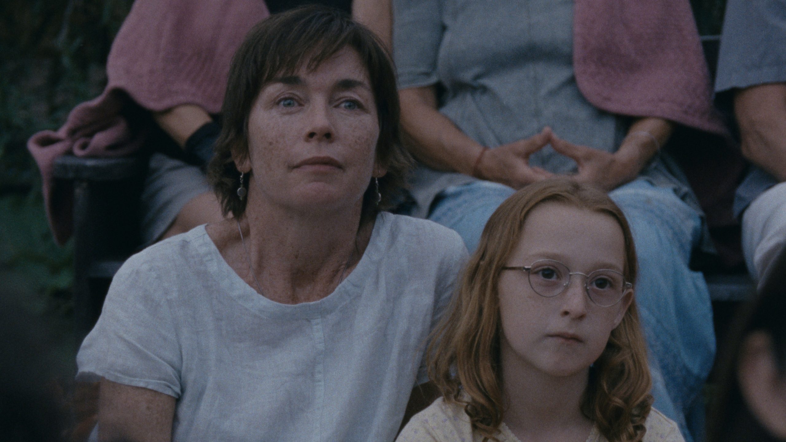 A woman in a white t-shirt and a young girl in glasses sit together on the grass.