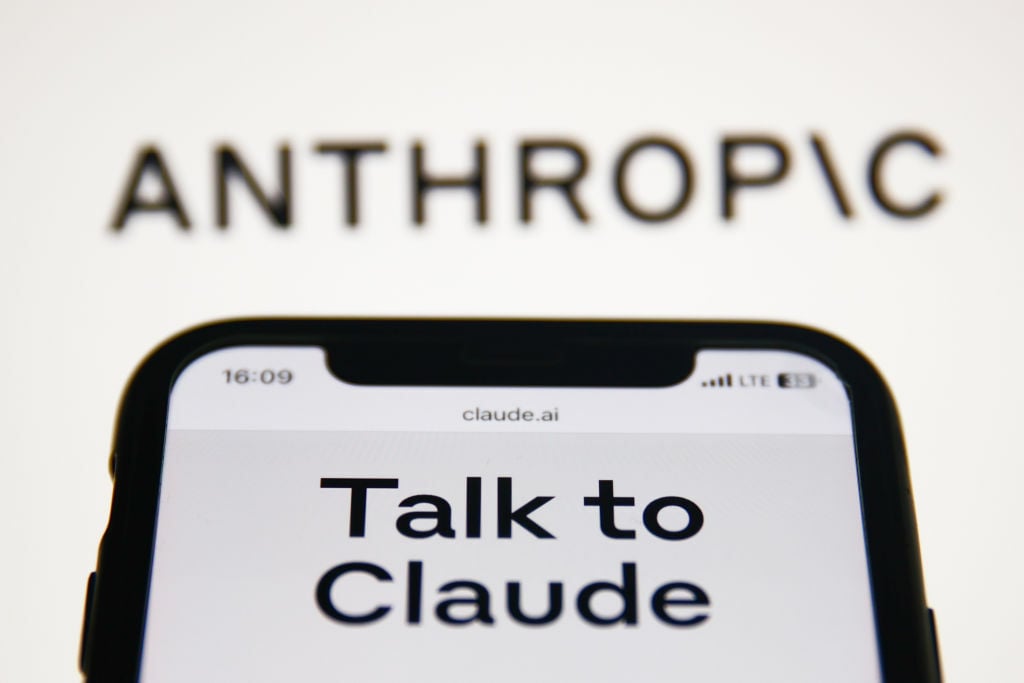 Anthropic Claude website displayed on a phone screen and Anthropic logo displayed on a screen in the background