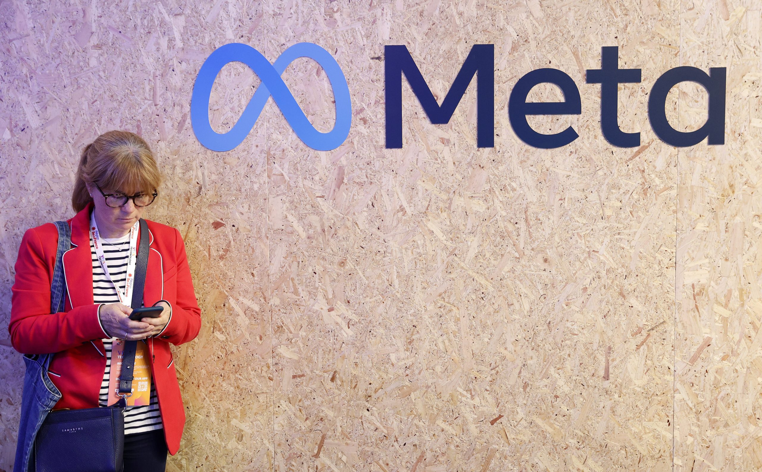 A woman in a red blazer leans against a wall with the Meta logo on it and looks down at her phone.