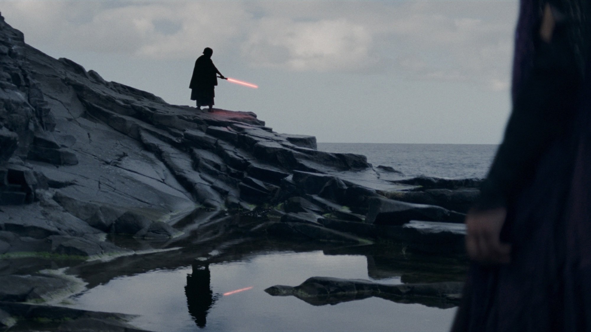 A figure in a dark cloak and helmet ignites a lightsaber while standing on a rocks near the ocean.