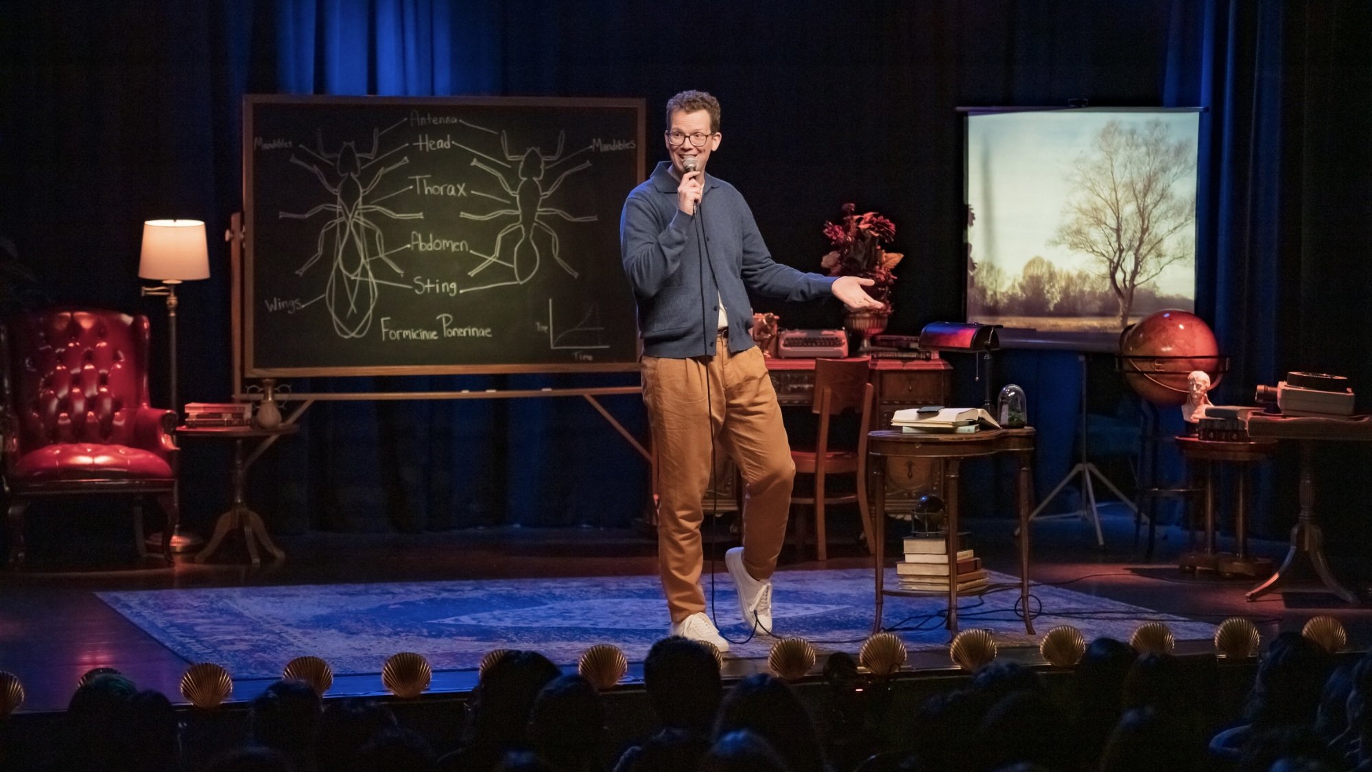 Hank Green performs stand-up onstage in front of a blackboard covered in anatomical drawings of ants.