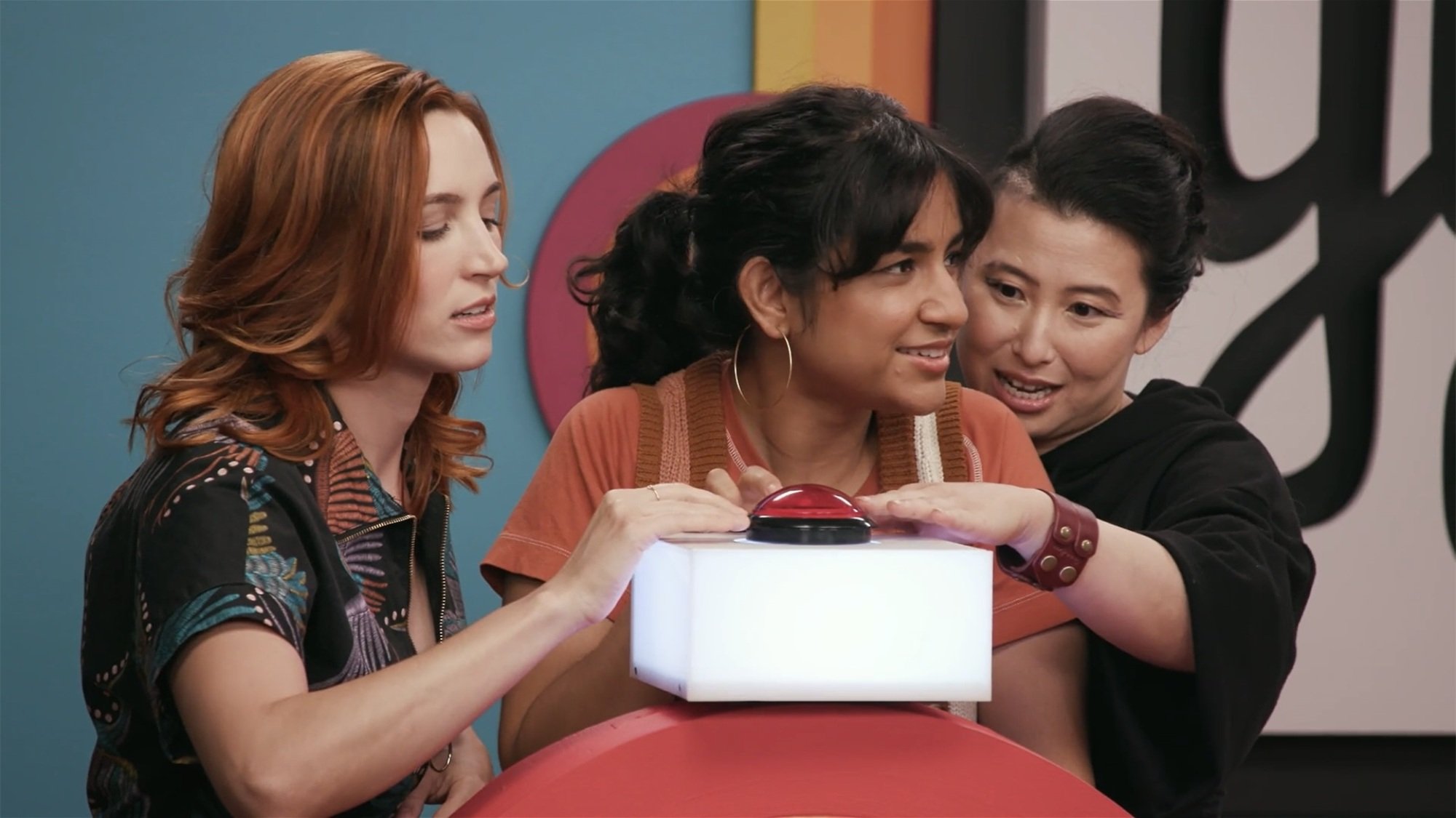 Three people gather around a game show podium with a red buzzer on top of it.