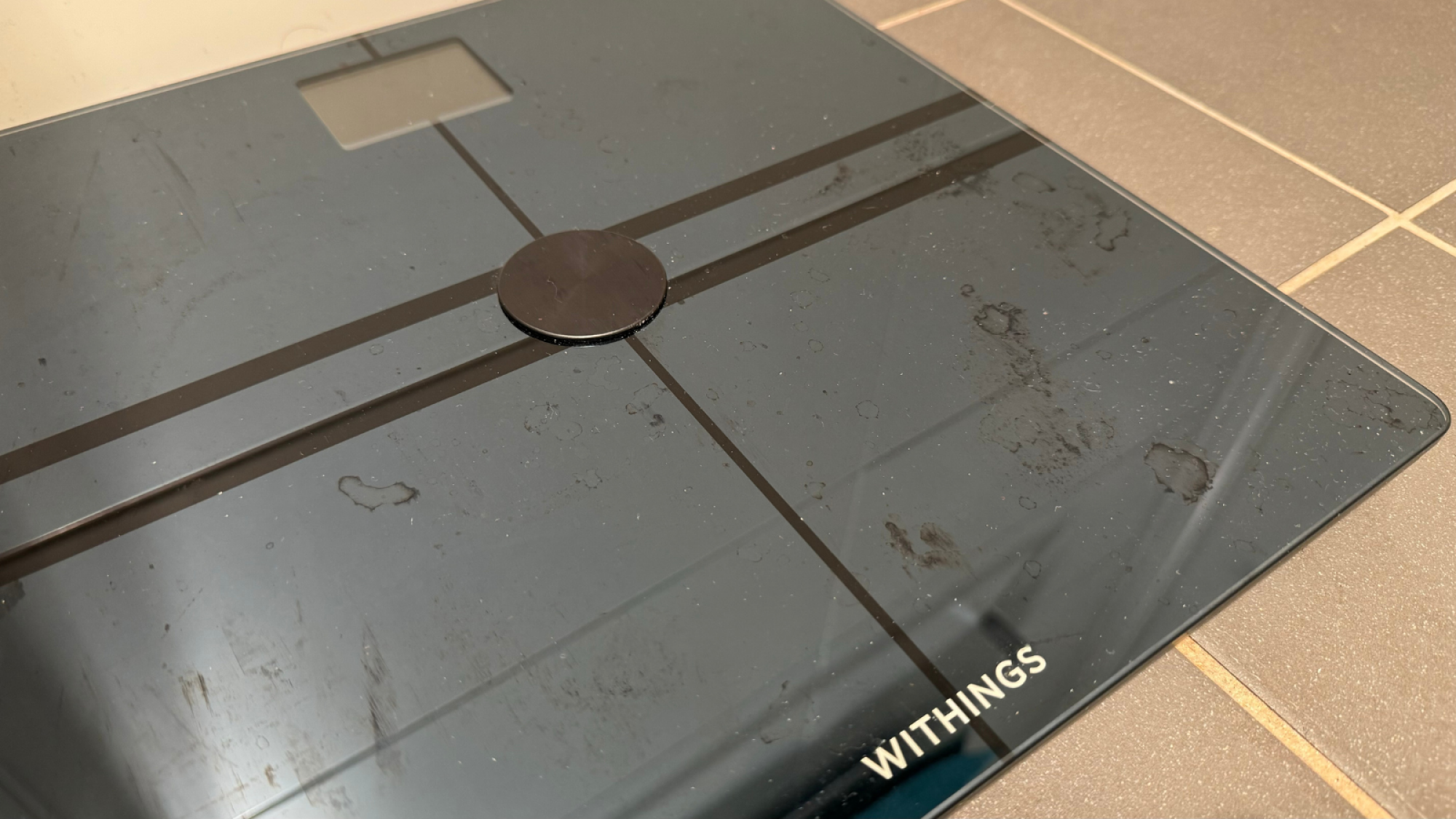 The Withings Body Comp smart scale on a tiled floor. It shows water stains.