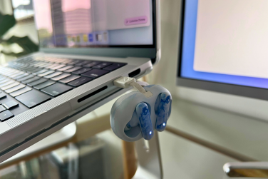 Skullcandy EcoBuds headphones charging on side of laptop with computer monitor and plant in background