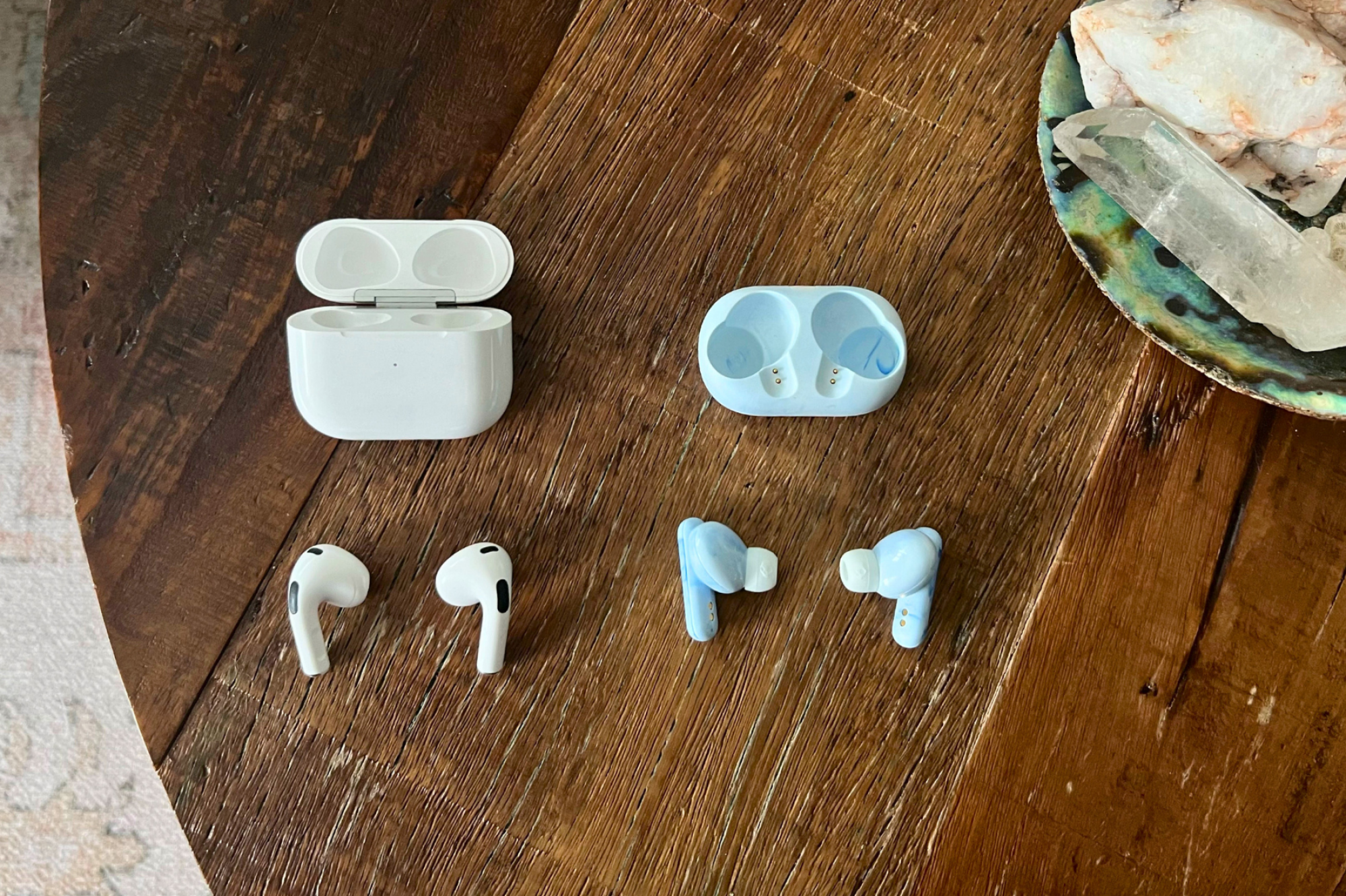 Apple AirPods and Skullcandy EcoBuds headphones and their cases laying on wood coffee table with seashell decor in peripheral