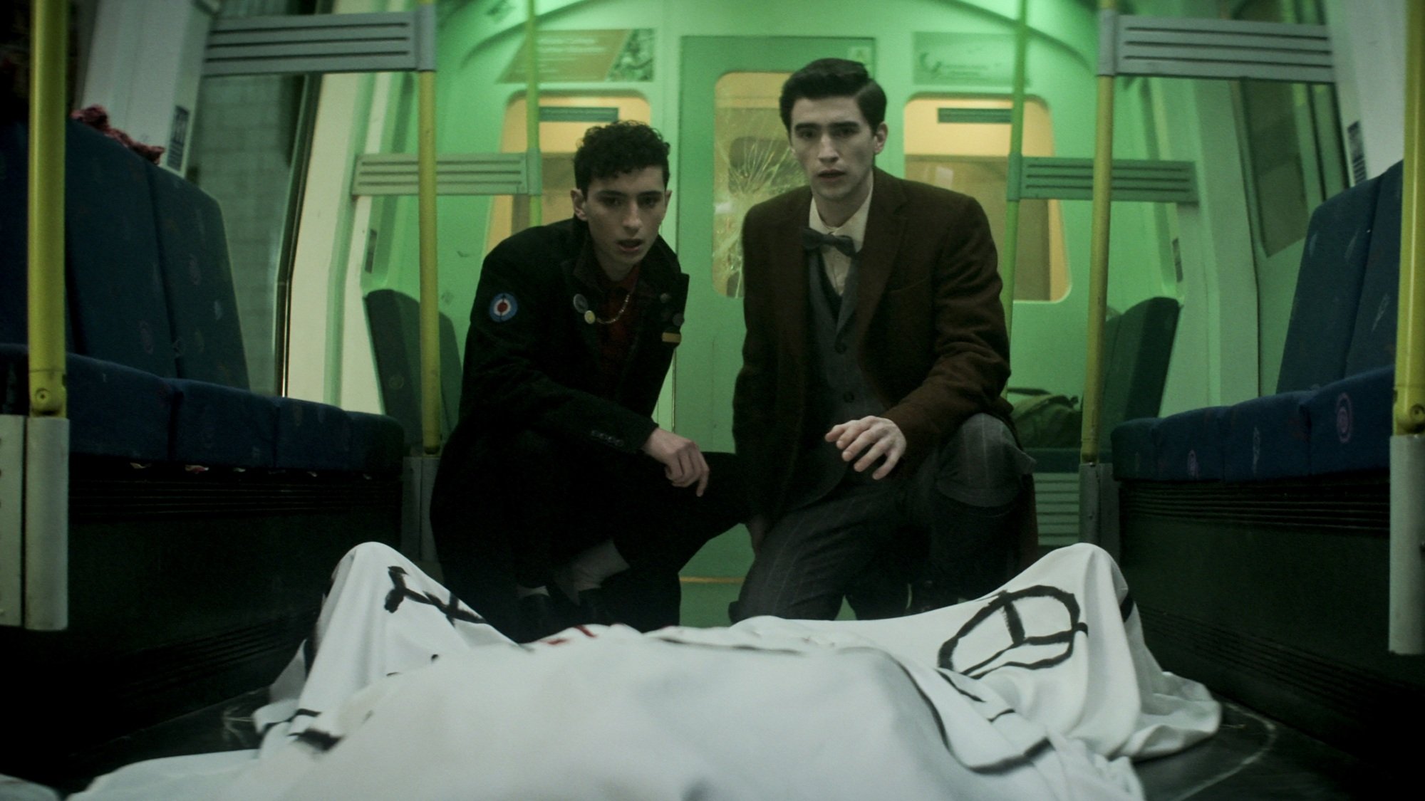 Two young men examine a covered dead body in a green-lit subway car.