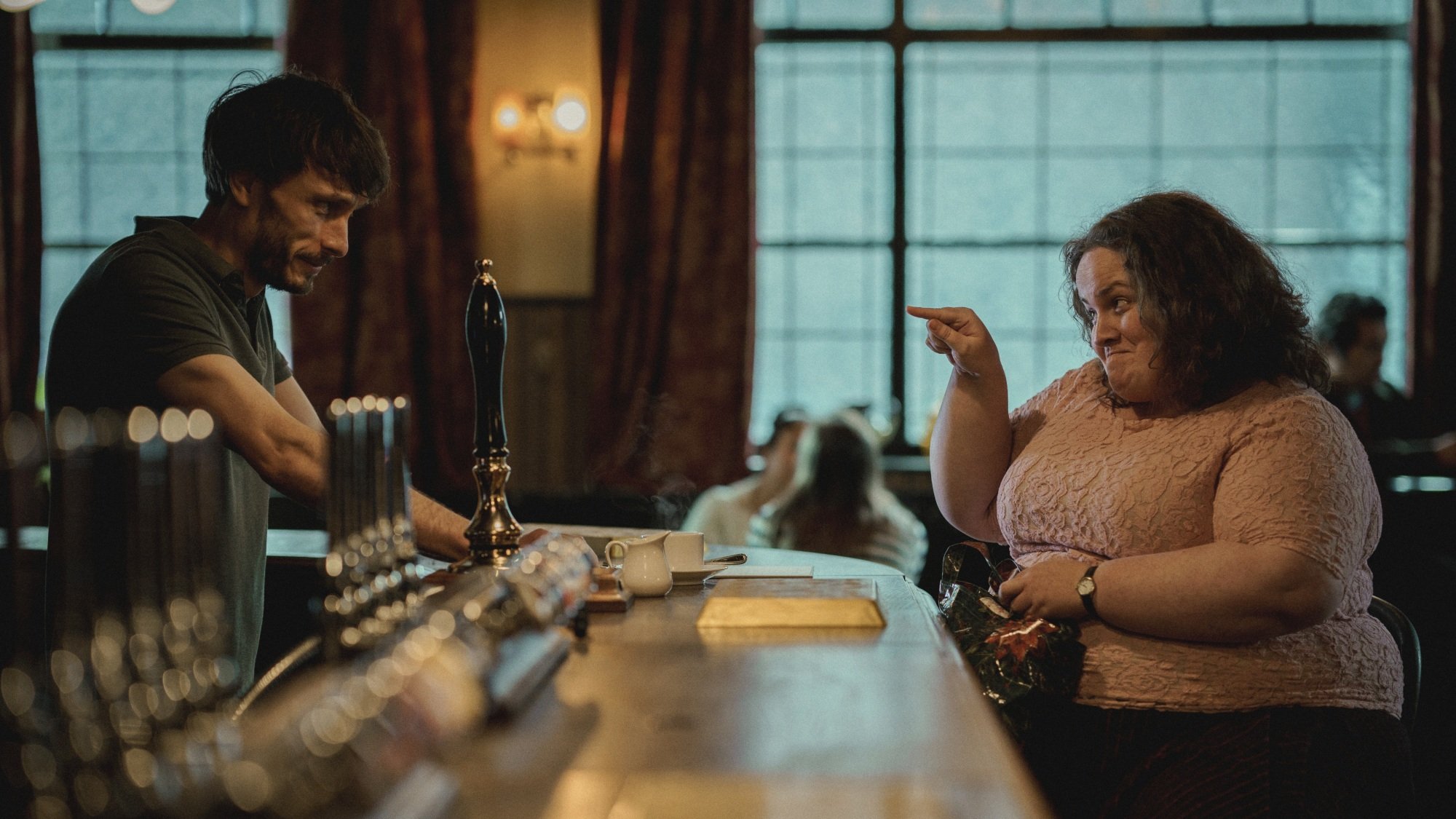 A woman in a pink shirt sits at a bar, pointing at the smiling man behind it.