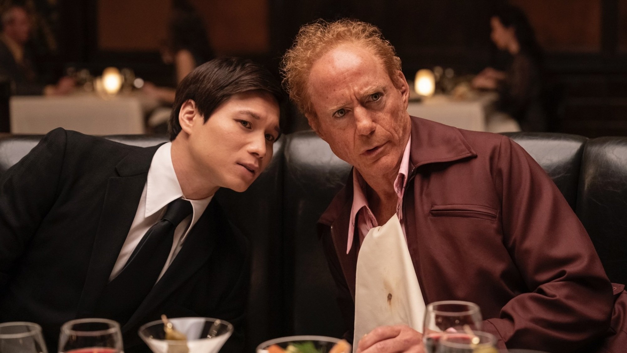 Two men seated next to each other in a restaurant booth lean in to speak to each other.