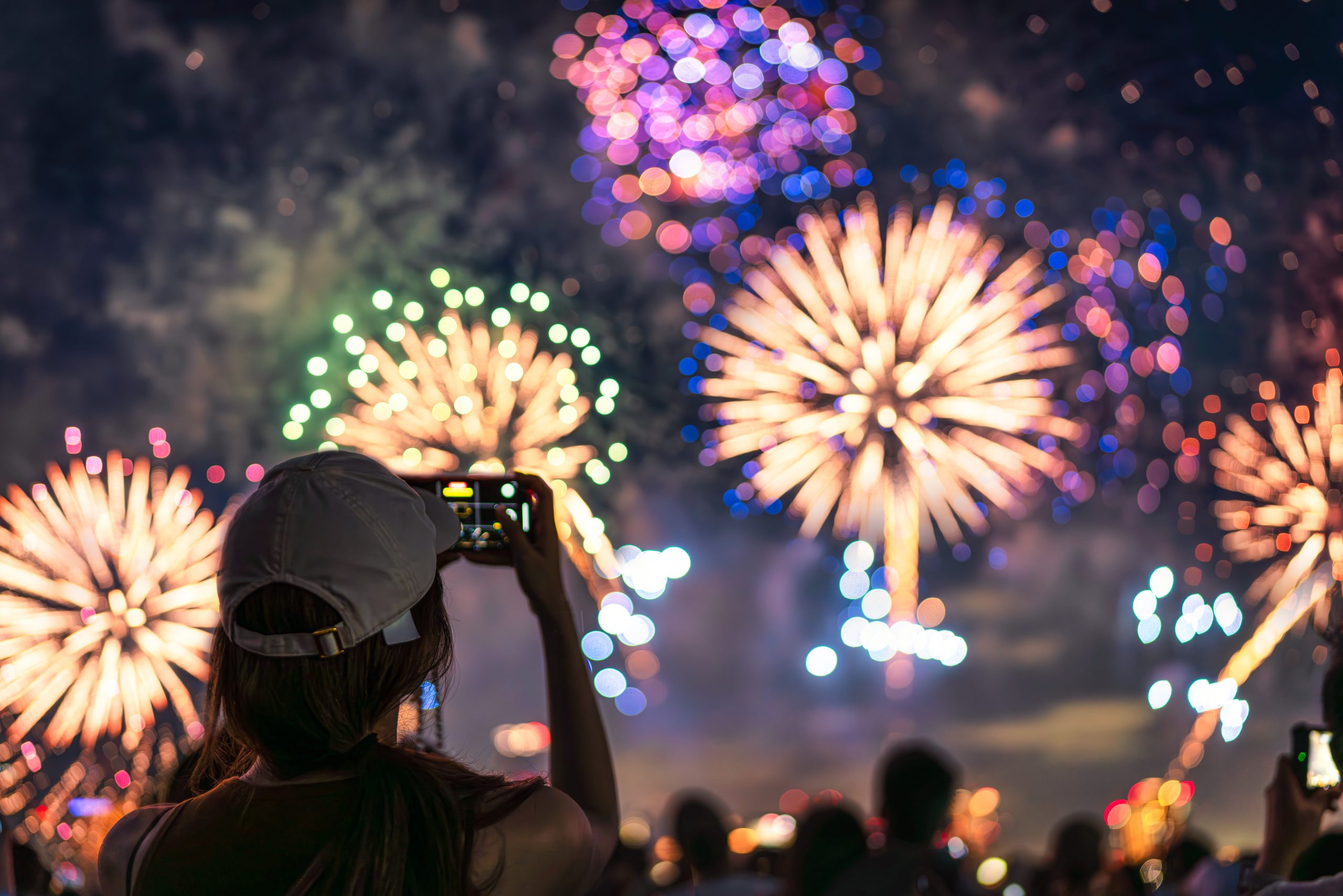 The back of a girl in a baseball cap filming a fireworks display on her mobile phone