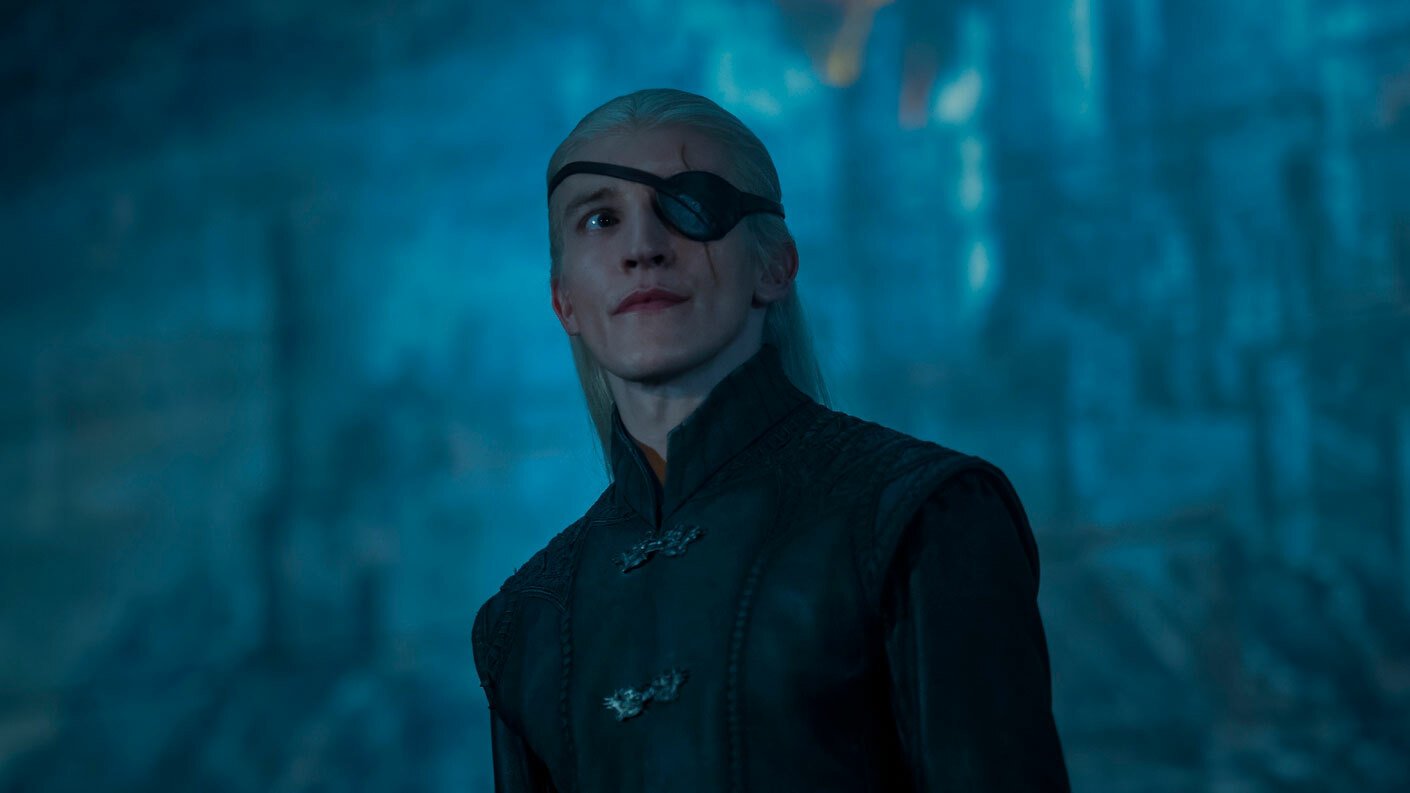 A man with blonde hair and an eyepatch stands in a gloomy room.