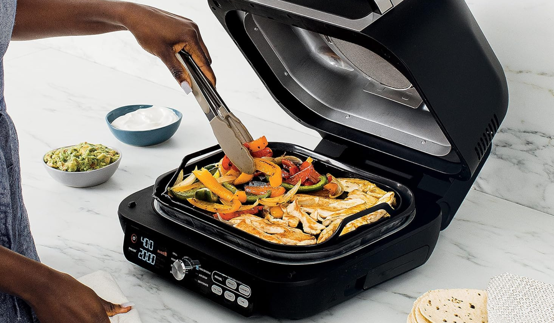 Person's hand using tongs to cook vegetables and chicken on Ninja griddle