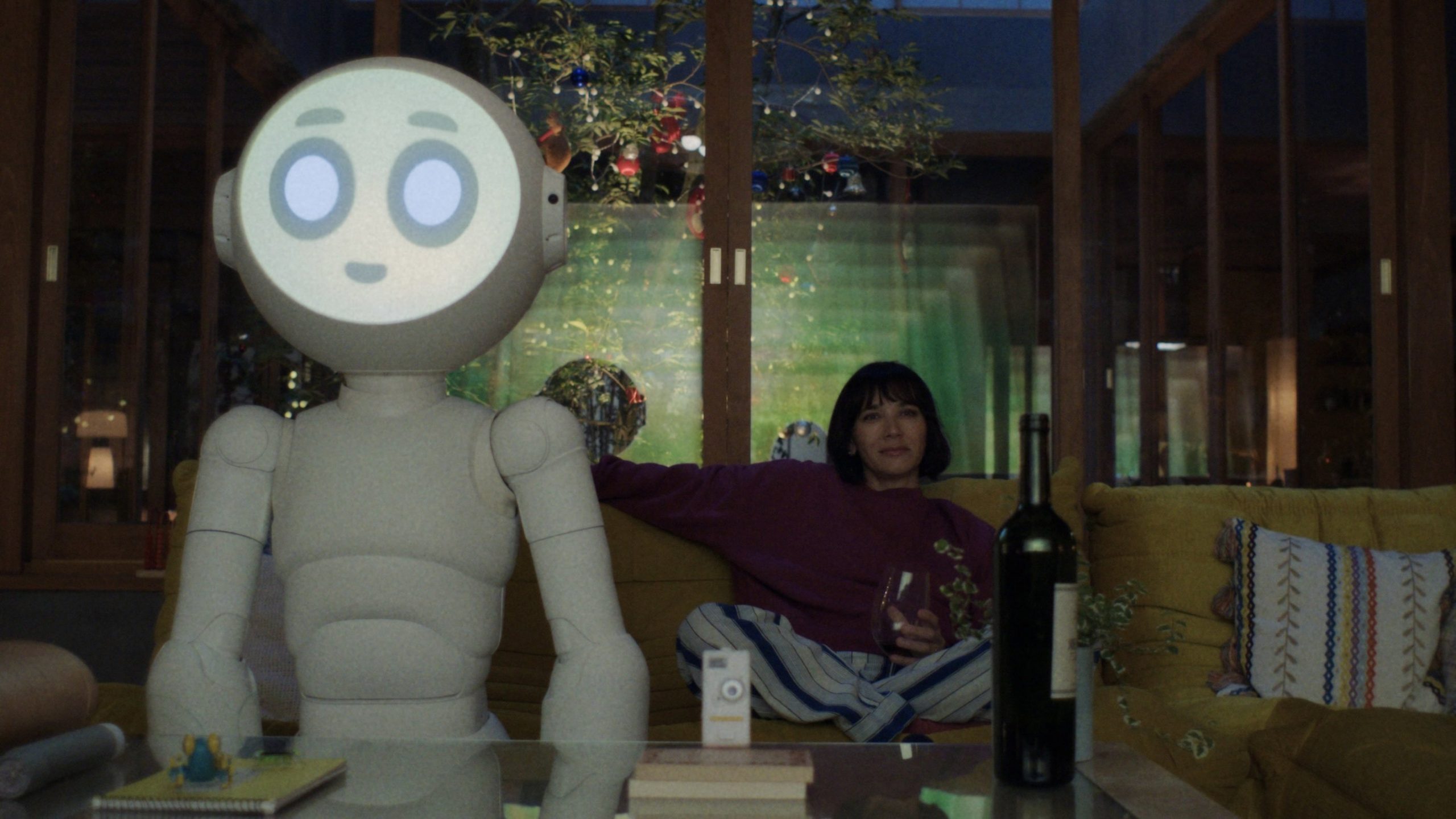 A large white robot with a glowing face and a human woman sit on a couch watching TV.