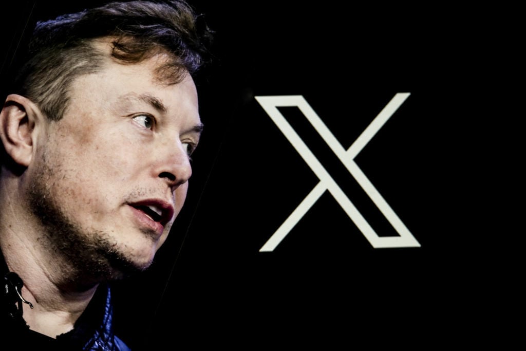 Elon Musk's photo is displayed on a phone screen in front of a computer screen displaying. the Twitter/X logo.