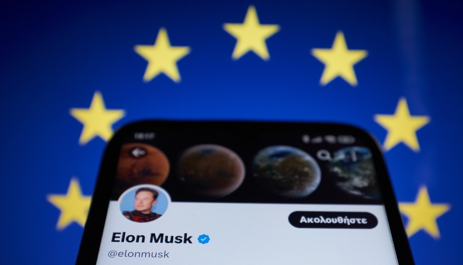 Elon Musk's bluecheck account on a phone against a European Union flag. 