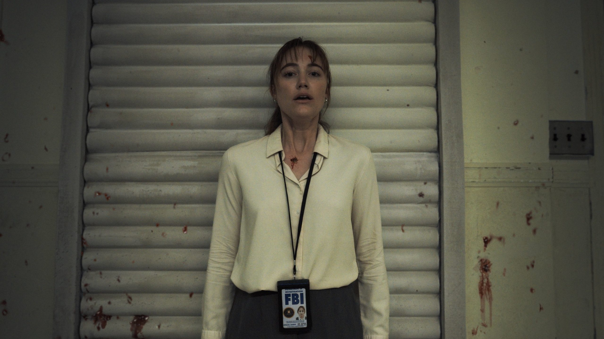 A woman in a blood-spattered white button-up shirt presses her back up against the wall of an interrogation room.