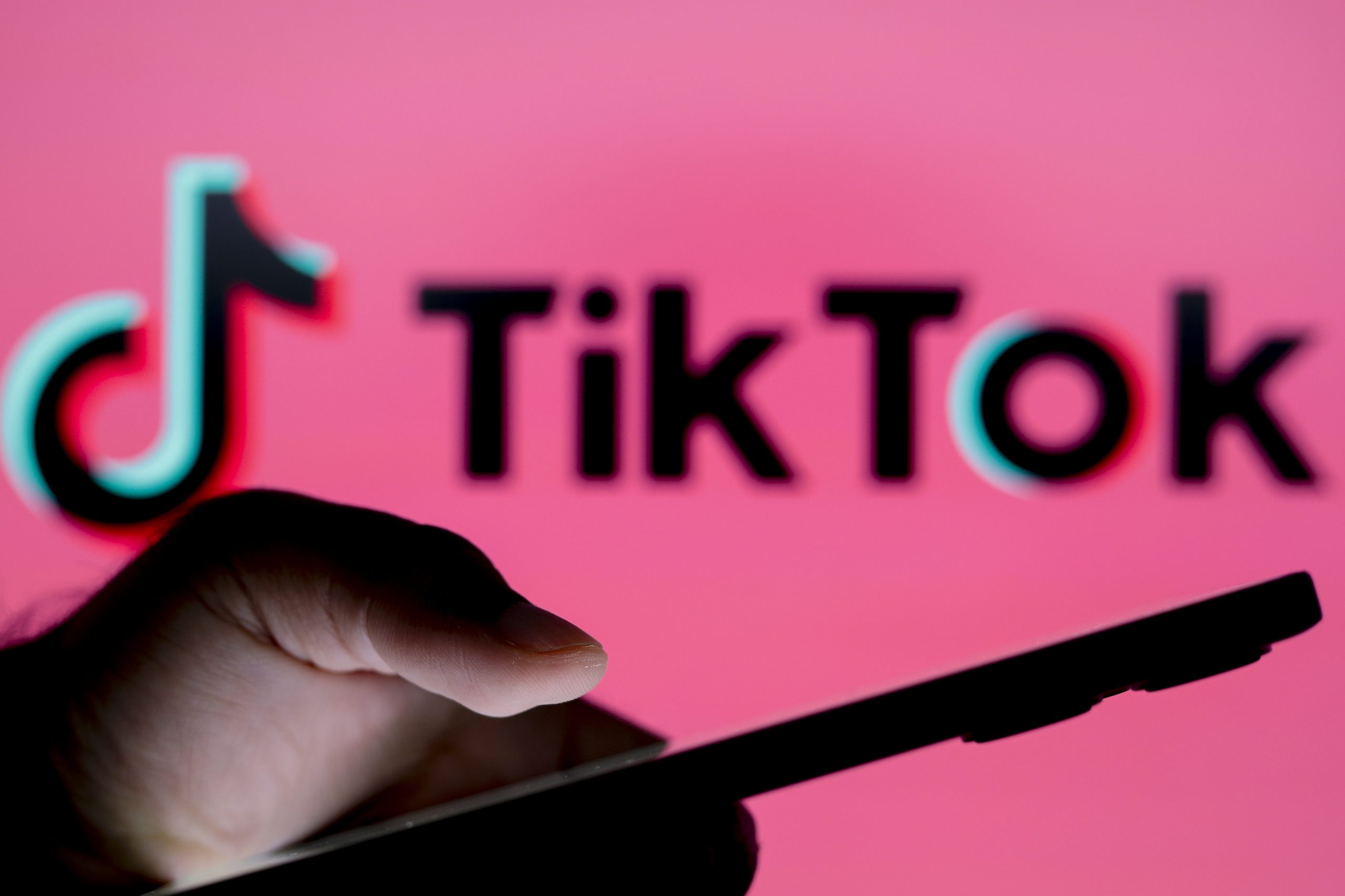 A hand holding a phone in front of a blurry sign reading TikTok on a pink backdrop.