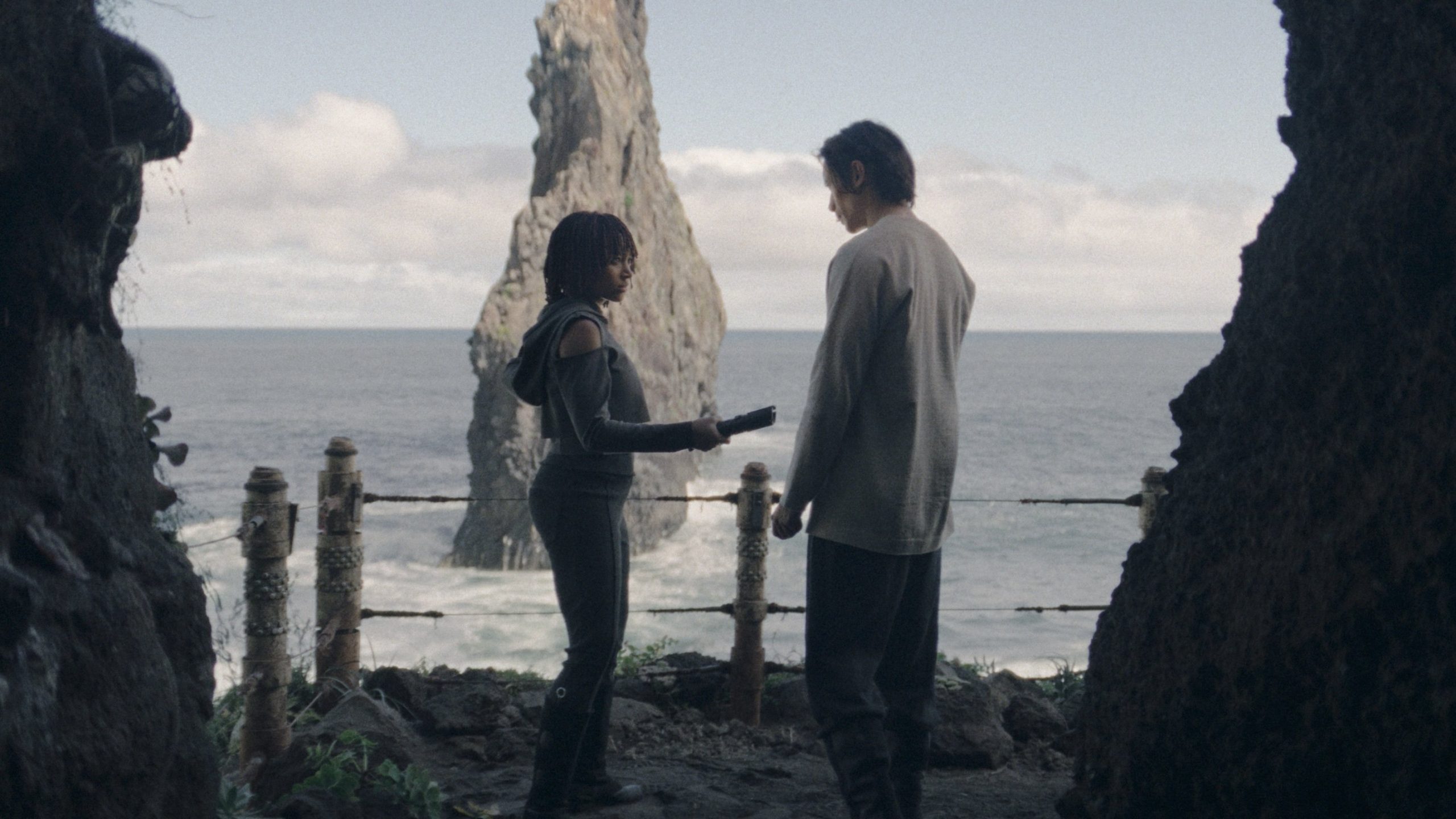 Osha holds an unlit lightsaber between her and the Stranger; the two stand at a cave entrance overlooking the ocean.