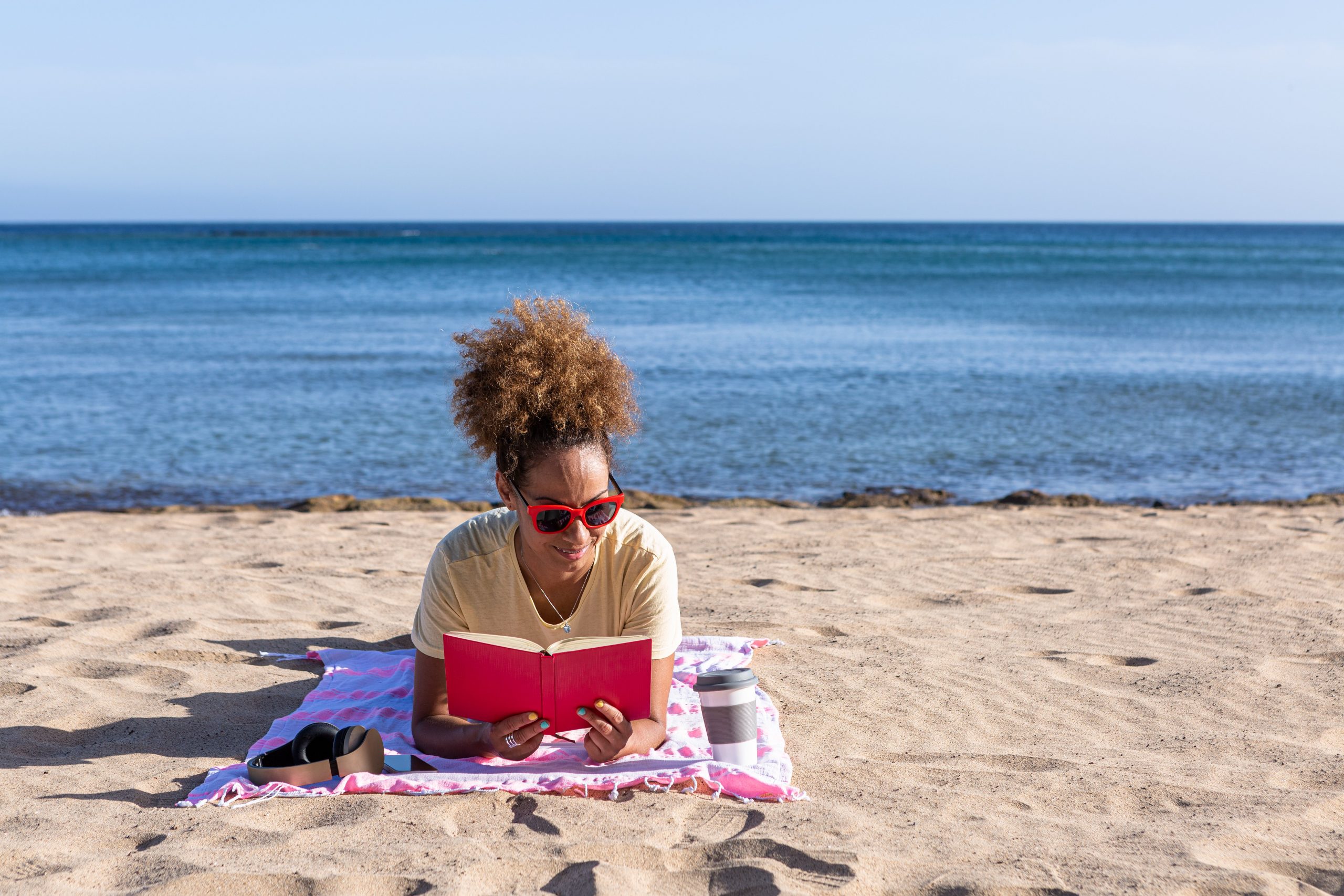 a person lays on a beach towel on the beach reading a red book with the ocean behind her