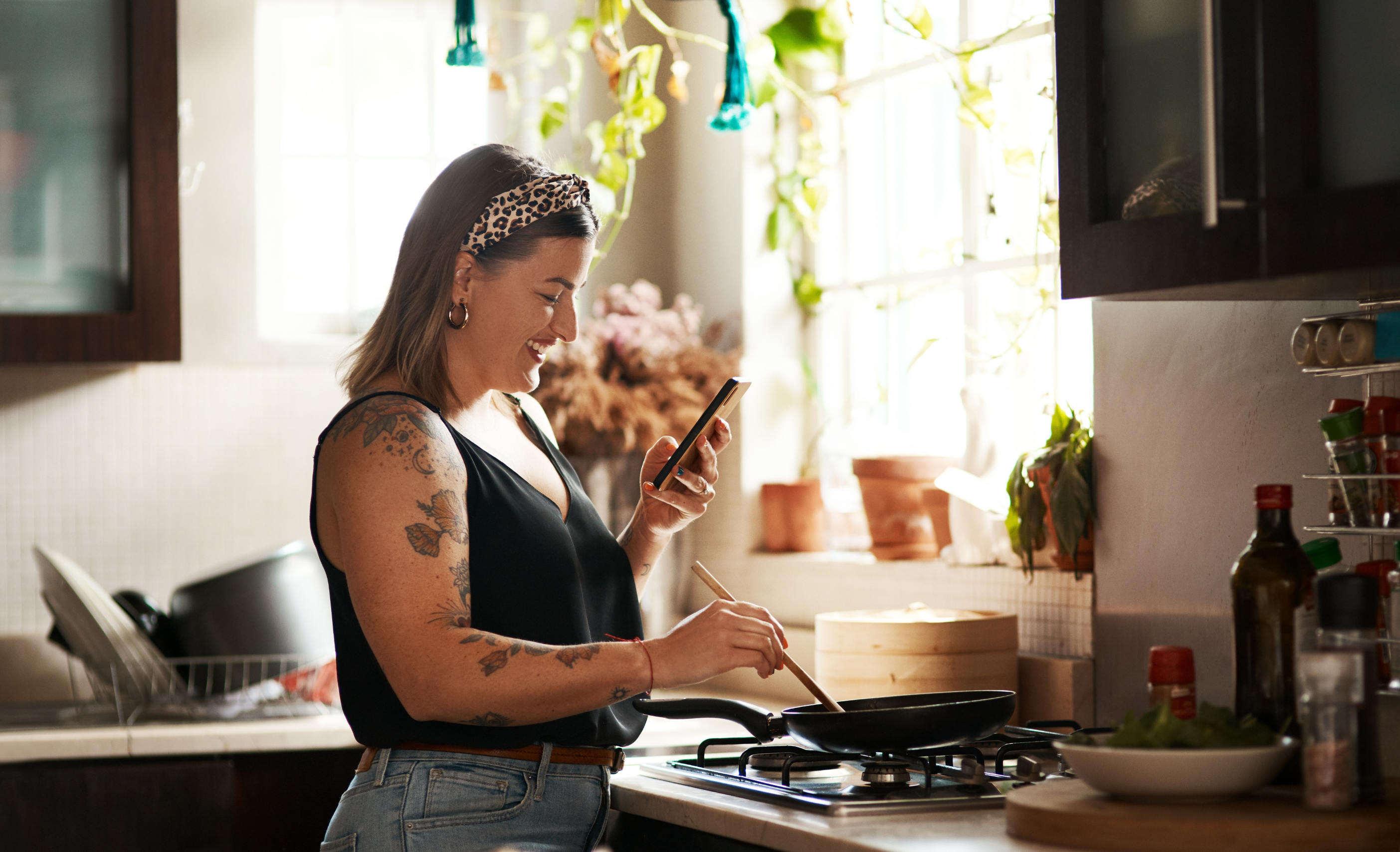 Happy, woman cooking food with smartphone and in kitchen of a home. Dinner or lunch, multitasking and plus size, natural person with tattoo on cellphone following recipe motivation for healthy diet