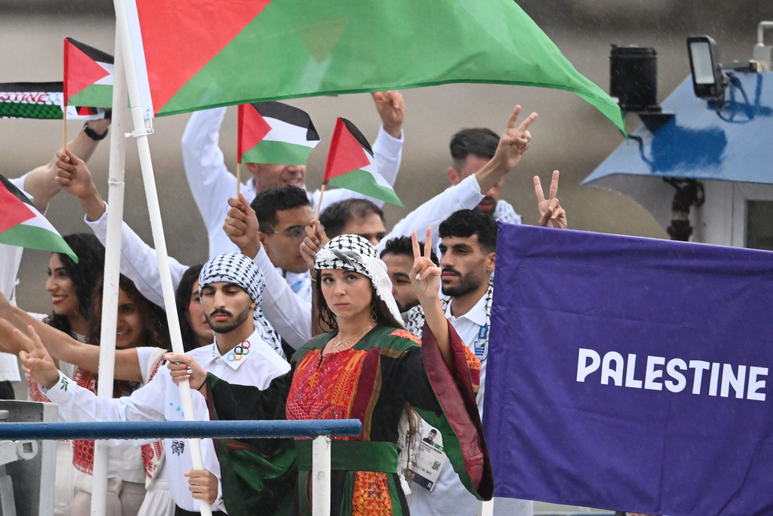 A group of Palestinian athletes hold up flags and peace signs while standing on a river barge.