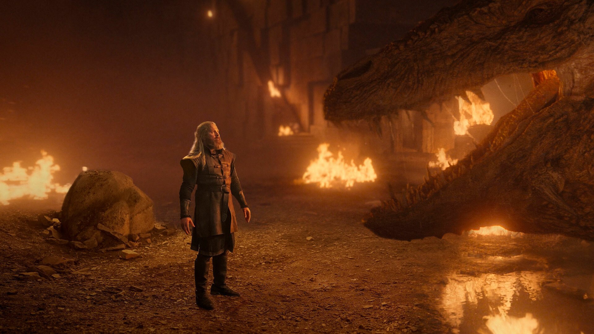 A bearded man stands in a gloomy cave, looking up at a dragon.