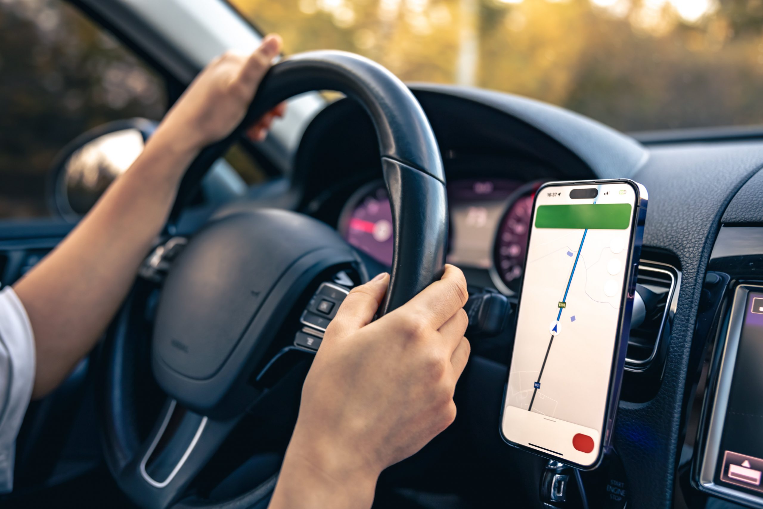 A person's hands on a car steering wheel. A phone with GPS navigation on the screen is attached to the dashboard.